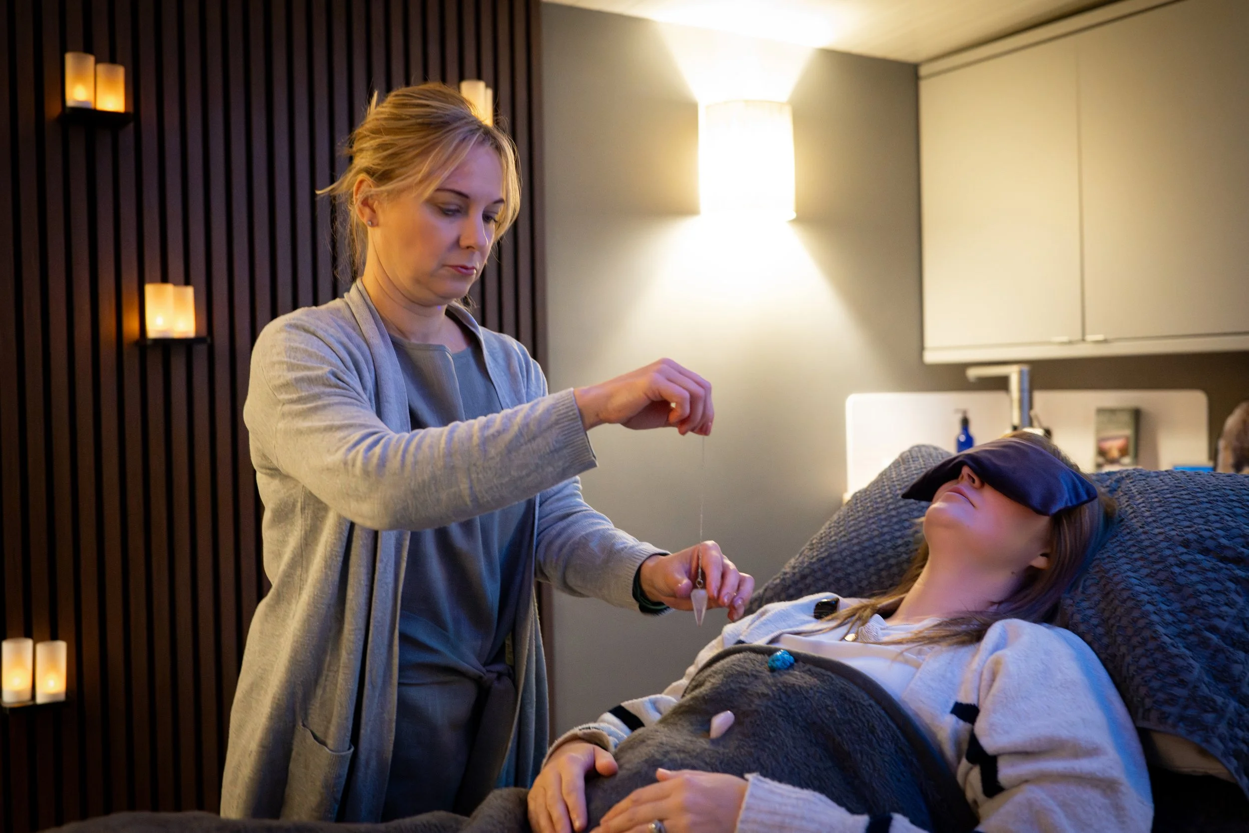 A woman with blonde hair, wearing a gray cardigan, is administering a liquid from a dropper into a young girl's mouth who is lying in bed with her eyes closed, blindfolded, and wearing a white sweatshirt. The room has dim lighting with candles on the