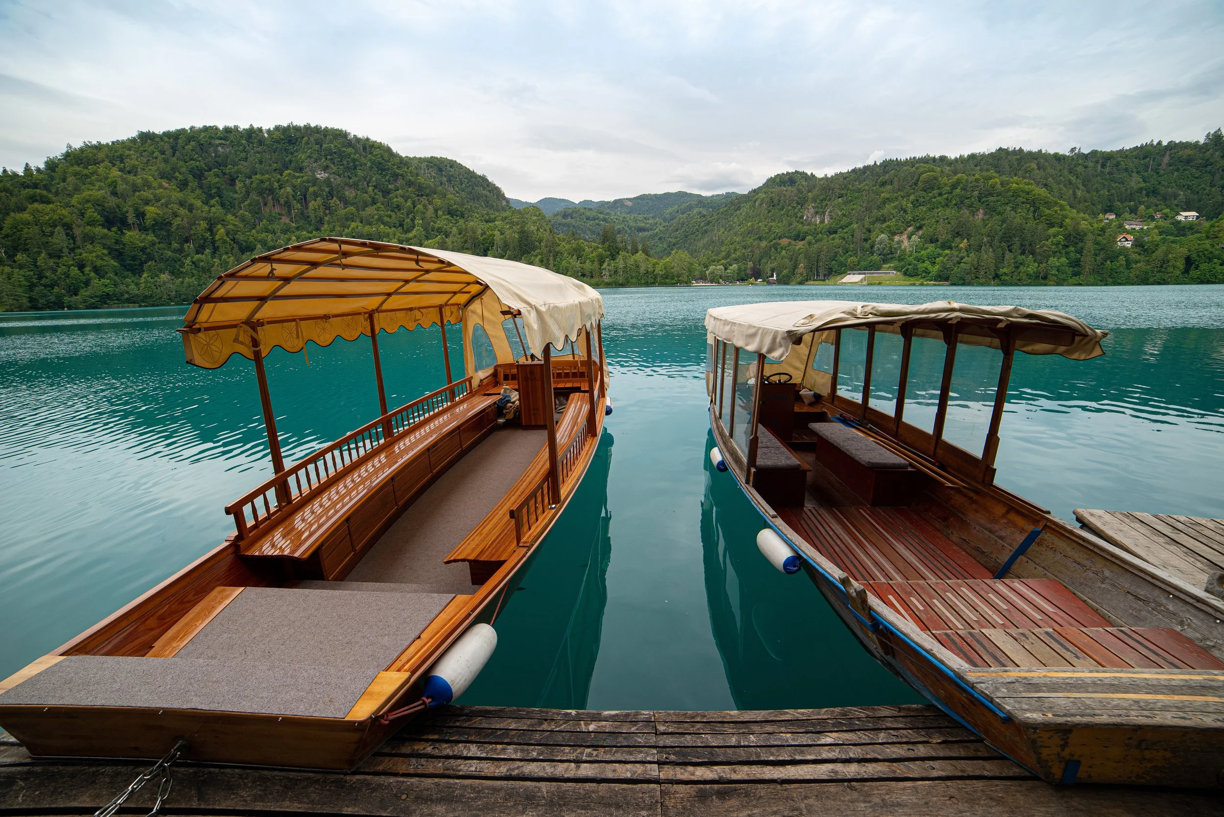 Two wooden boats with canopies docked at a pier on a lake, surrounded by green mountains and cloudy sky.