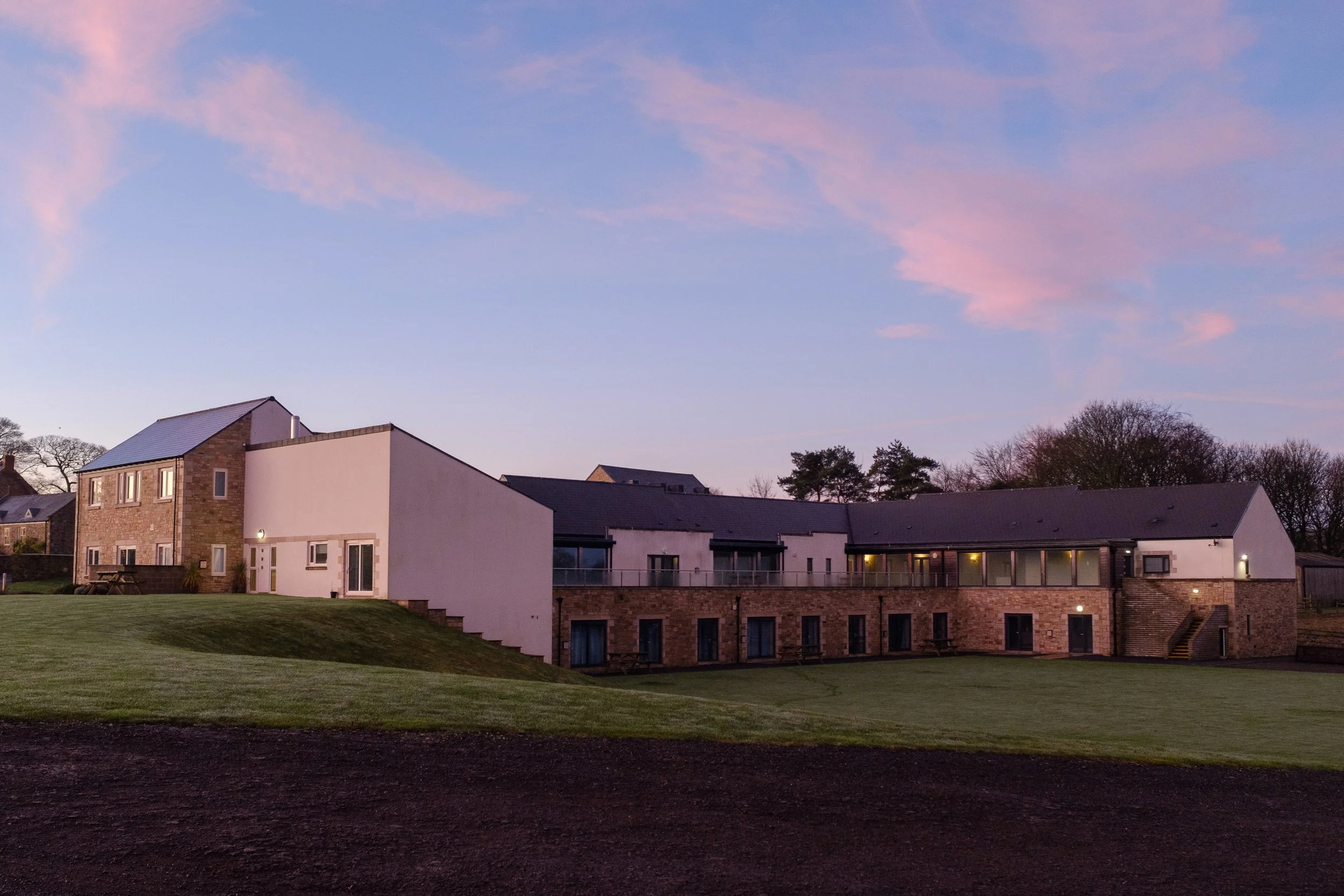 A modern multi-story residential building with a combination of stone and white exterior walls, large windows, and a sloped roof, set against a sky with pink and blue clouds at dusk, with a grassy yard in the foreground.