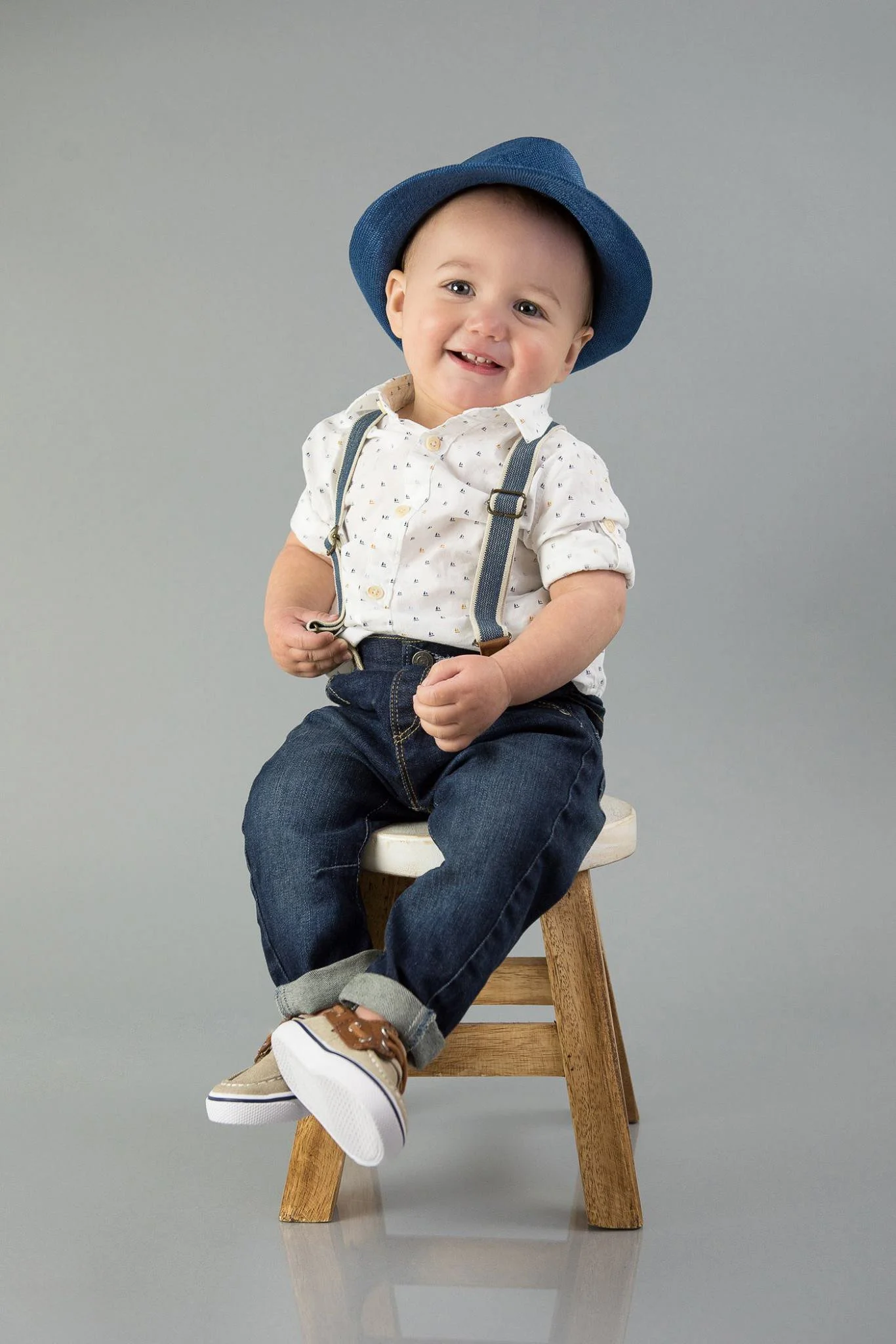 A smiling toddler boy sitting on a small wooden stool, wearing a blue hat, white shirt with small patterns, dark jeans with rolled-up cuffs, and brown sneakers.