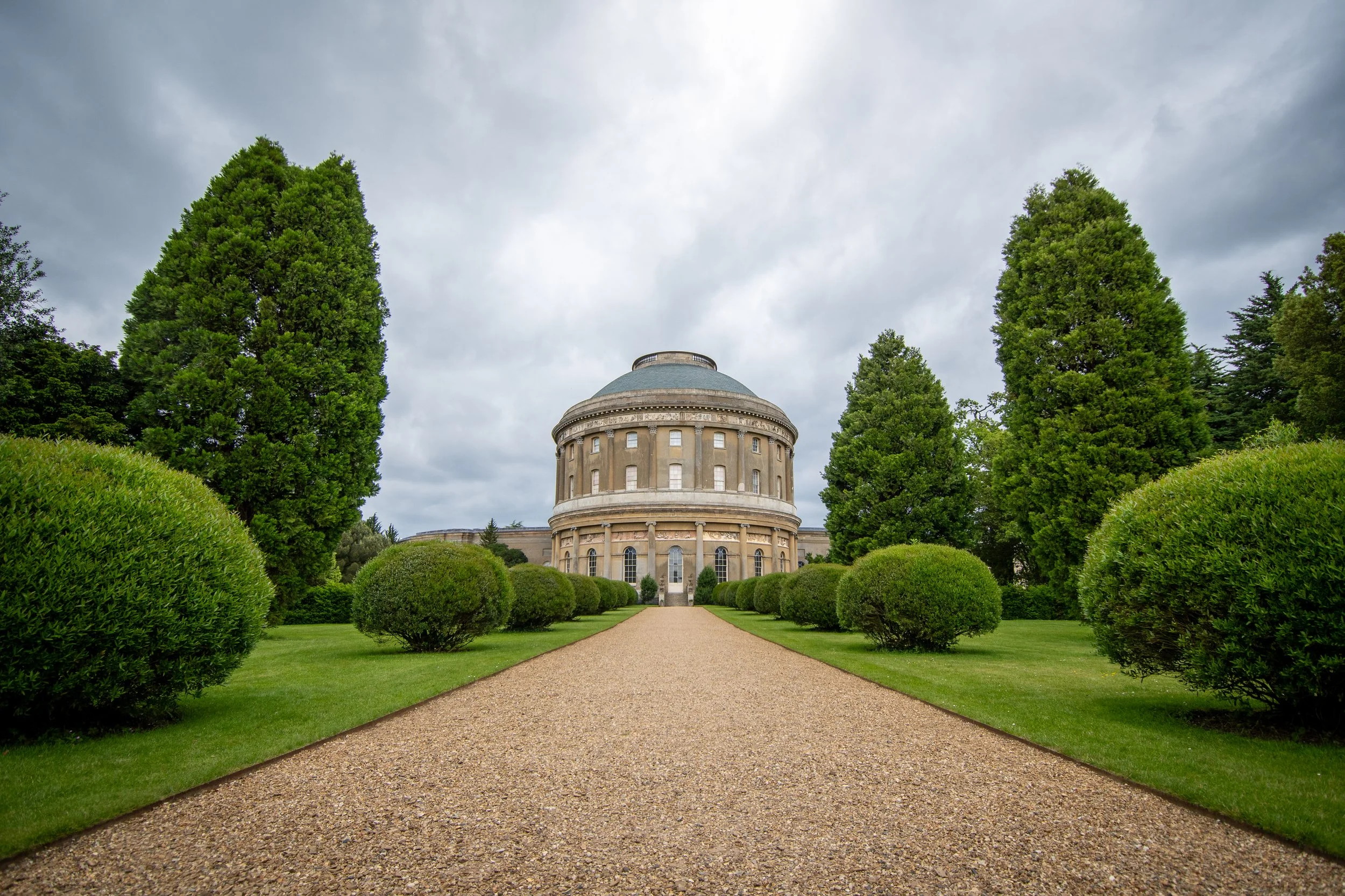 A large, historic circular building with a domed roof, surrounded by well-manicured bushes and tall trees, under a cloudy sky.