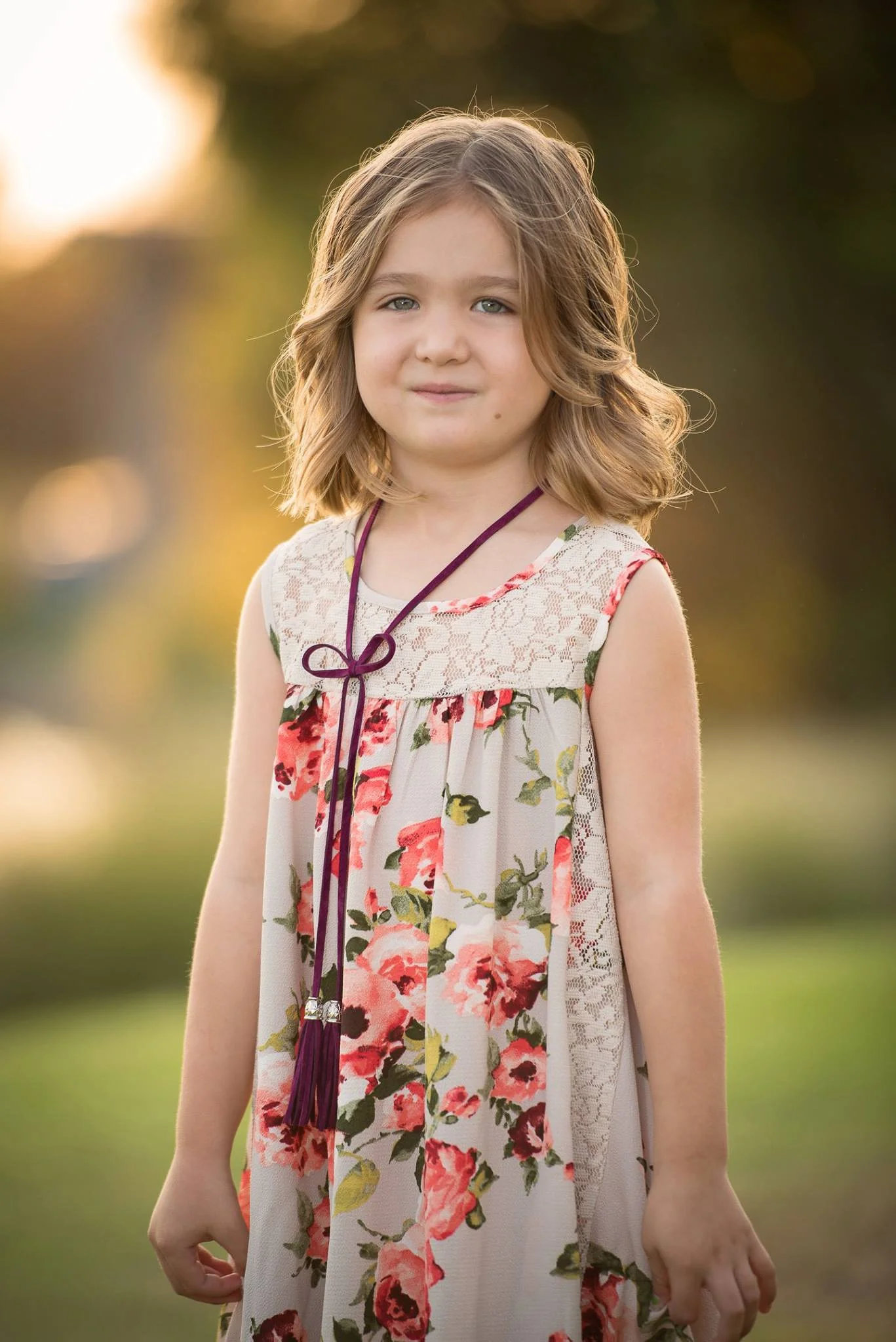 A young girl with shoulder-length wavy light brown hair, standing outdoors in the late afternoon or early evening with blurred trees and sunlight in the background. She is wearing a floral dress with lace details and a purple necklace.