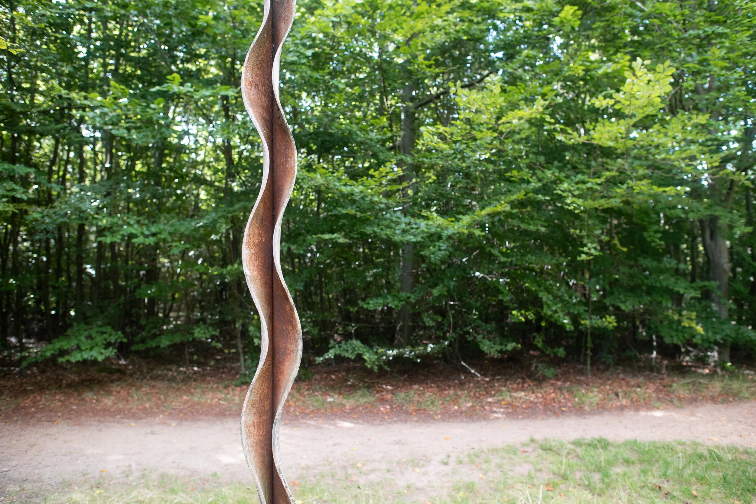 Close-up of a twisted wooden sculpture outdoors with a background of green trees and a dirt path.