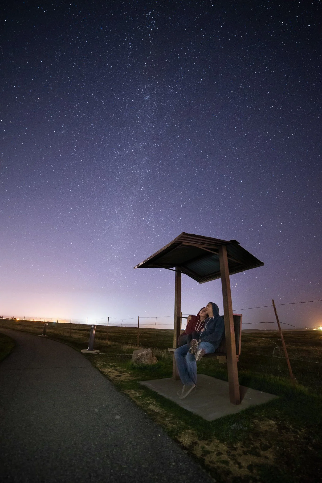 Two people sitting under a wooden shelter by a paved path, looking up at a starry night sky with the Milky Way visible.