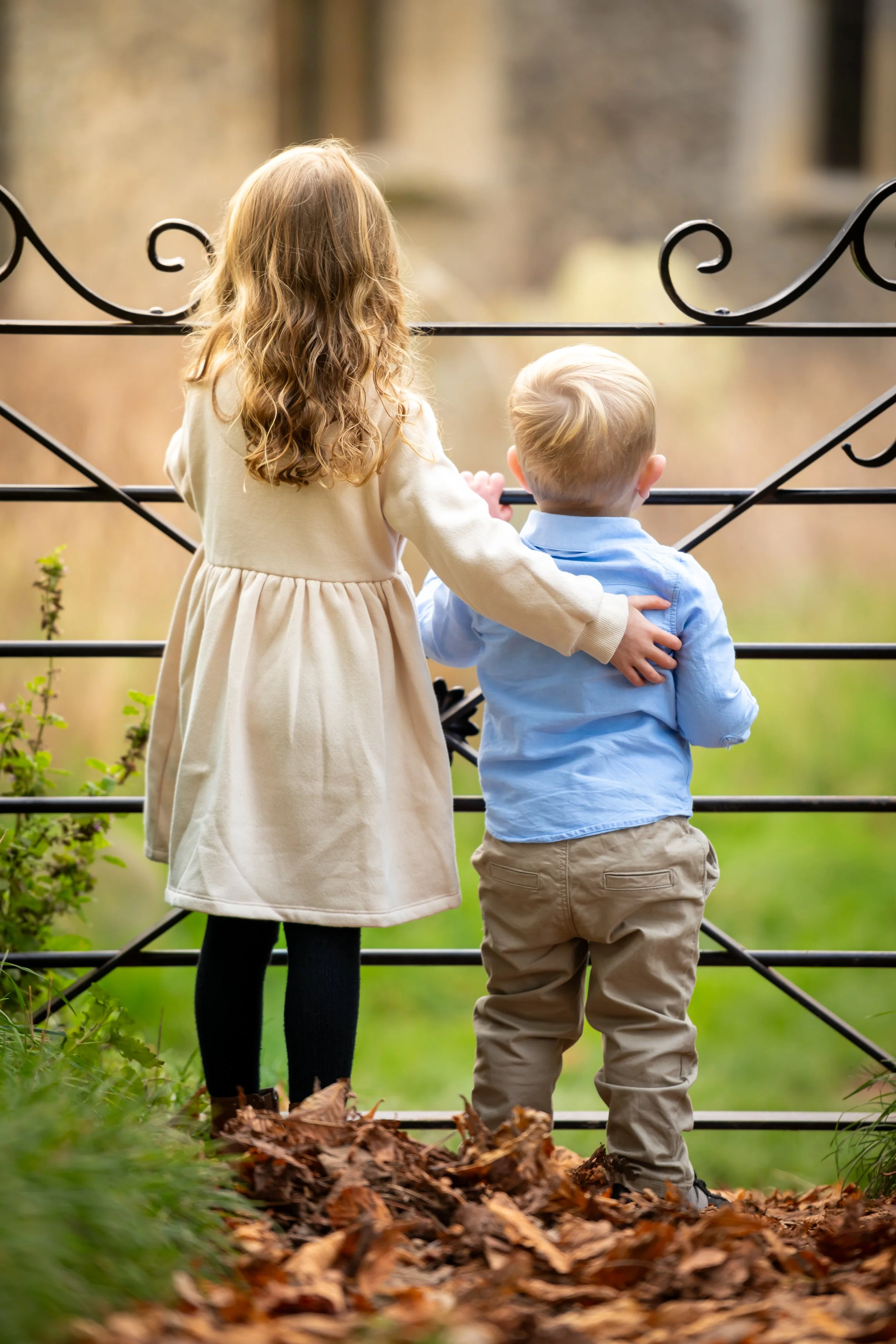 A young girl and boy standing together near a decorative metal gate, looking away outdoors in a natural setting with fallen leaves and green grass.