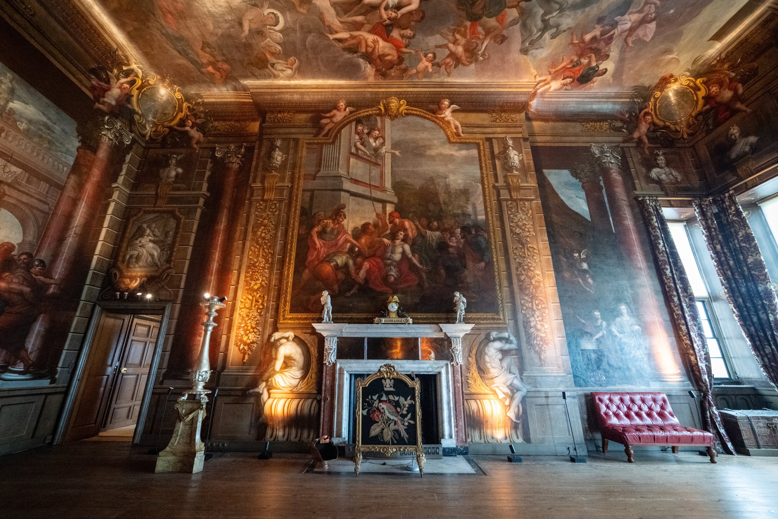 Historic room with ornate wood paneling, large classical oil painting above fireplace, sculptures, and decorative drapes.