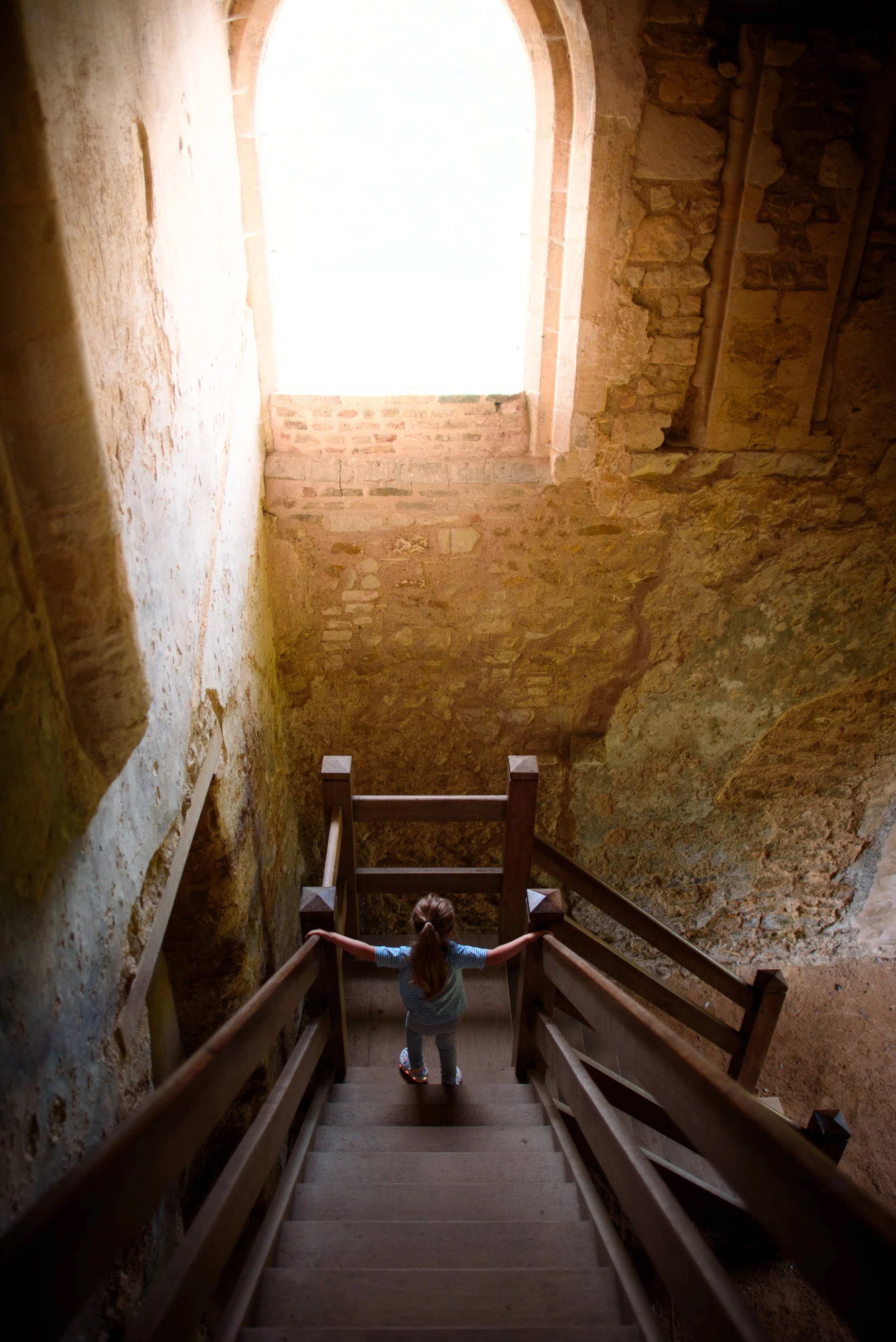 A young girl with long hair in a ponytail wearing a striped shirt and jeans holding onto the wooden handrails of a staircase inside an old stone building, with a large window letting in sunlight.