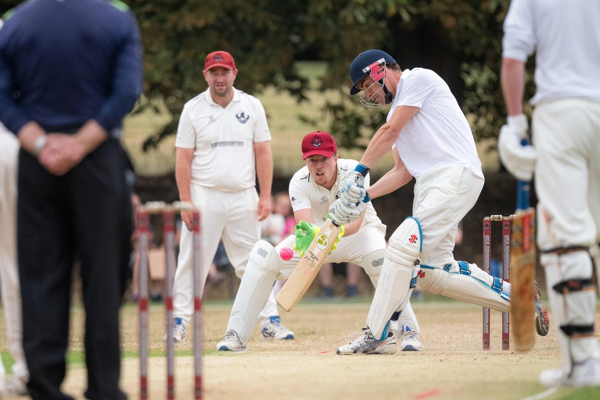 Cricket players in white uniforms and helmets during a game, with an umpire and other players visible in the background.
