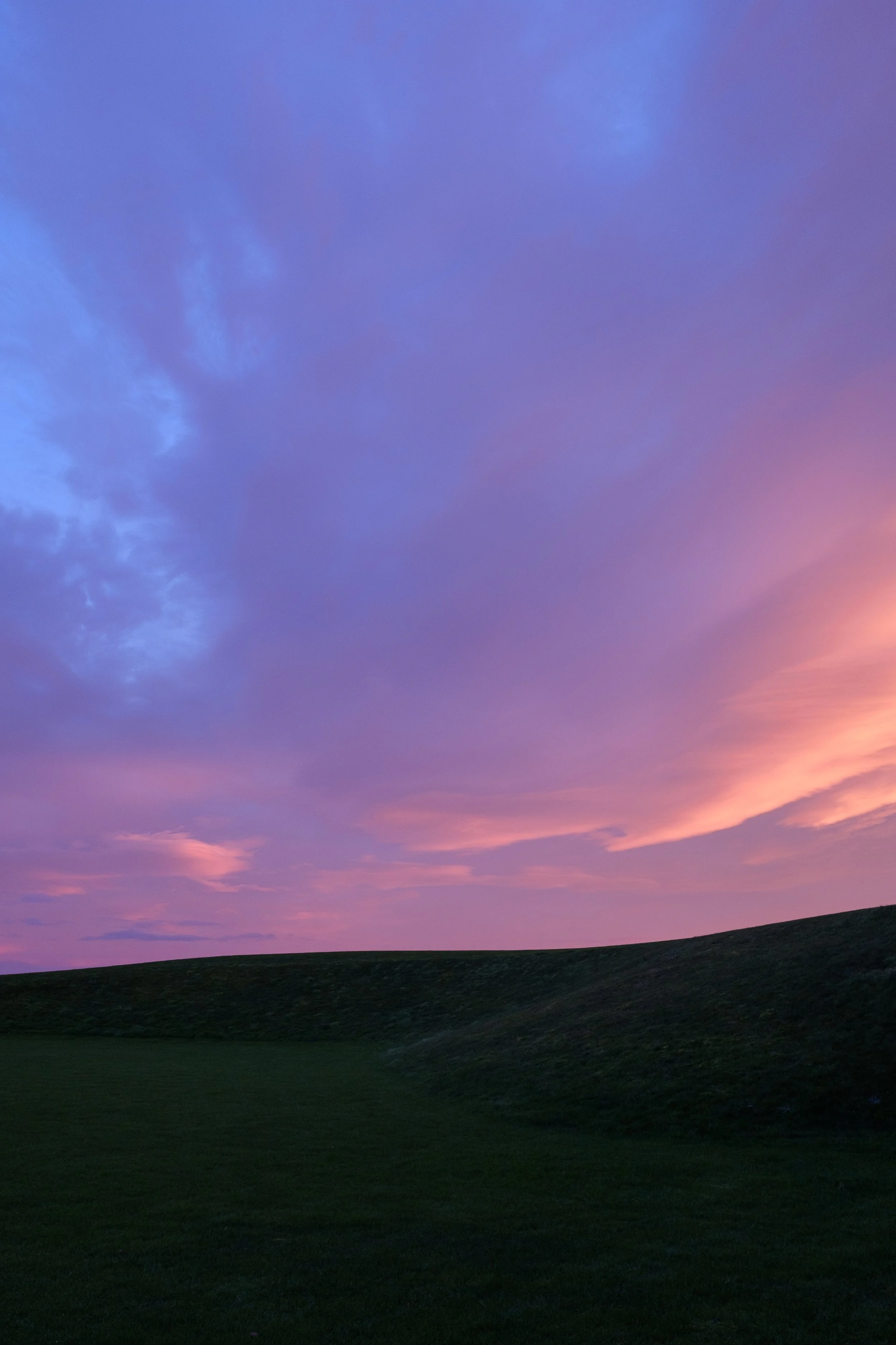 A sunset or sunrise sky with shades of pink, purple, and blue over a dark grassy hill.