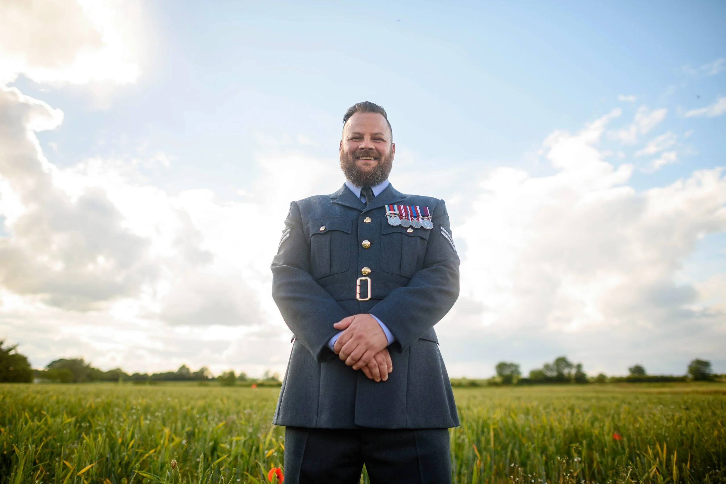 A man in a military uniform standing in a grassy field with a partly cloudy sky behind him.