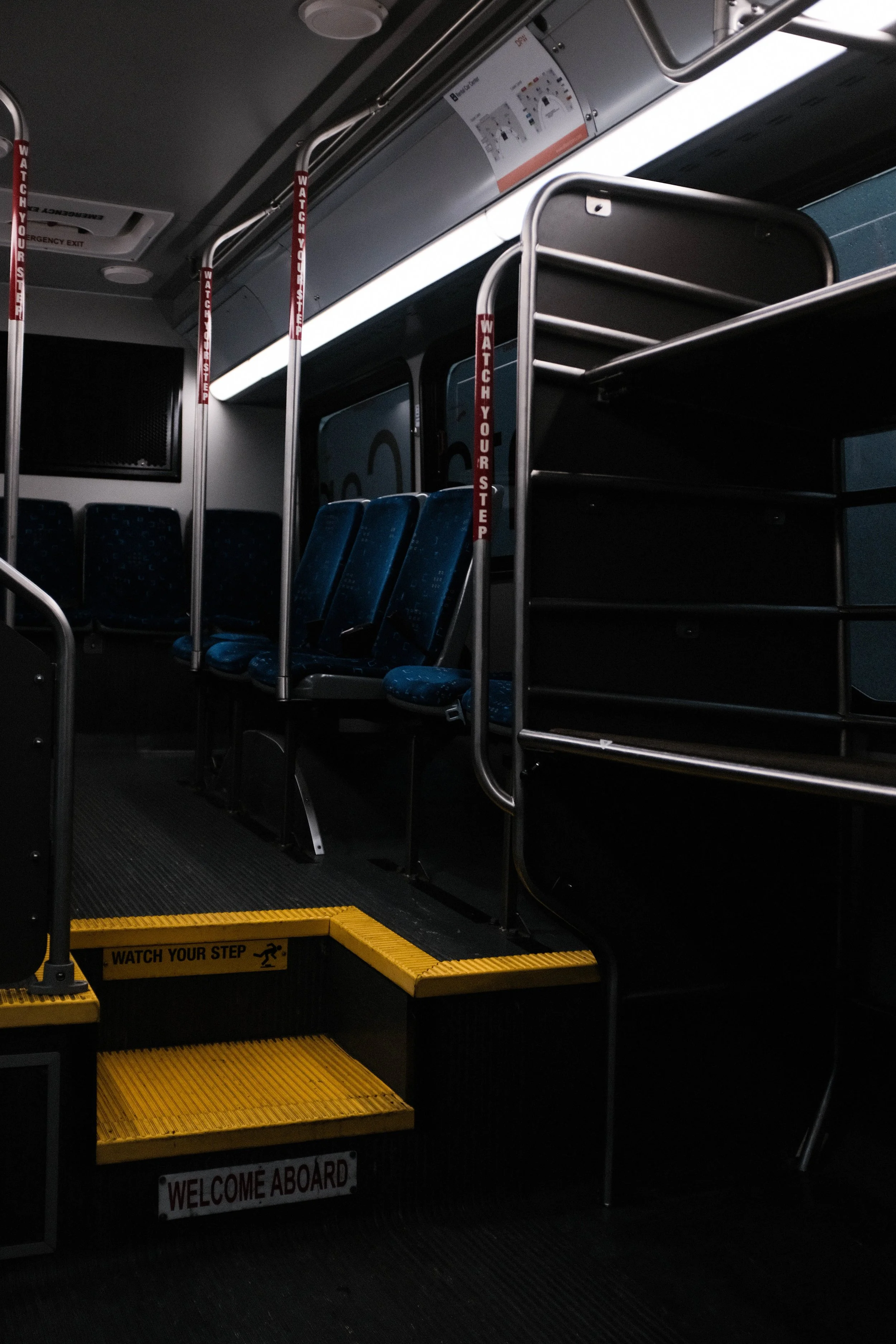 Interior of a bus with empty blue seats, a step with yellow tape and caution signs, and safety bars with warning labels.