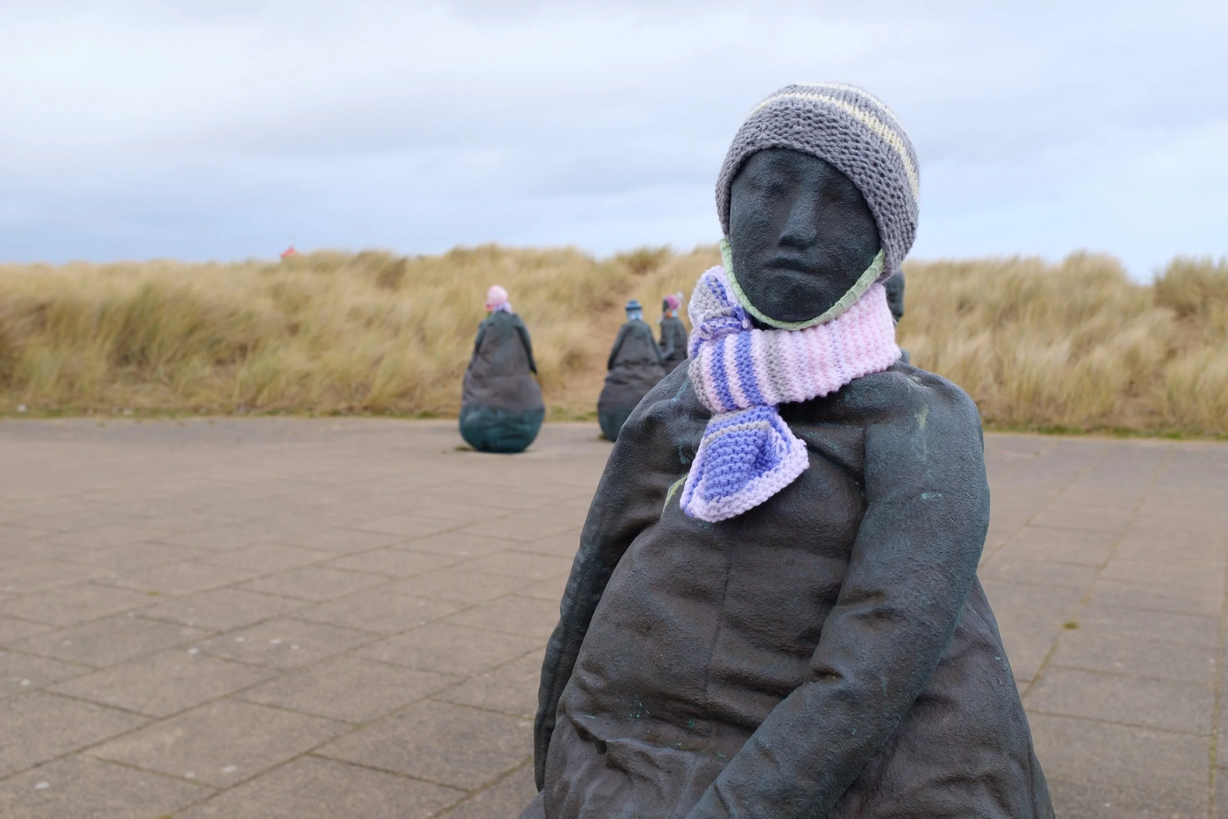 Sculpture of a seated figure with a knitted hat and scarf, with a dark face, in an outdoor setting with grassy dunes and other similar sculptures in the background.