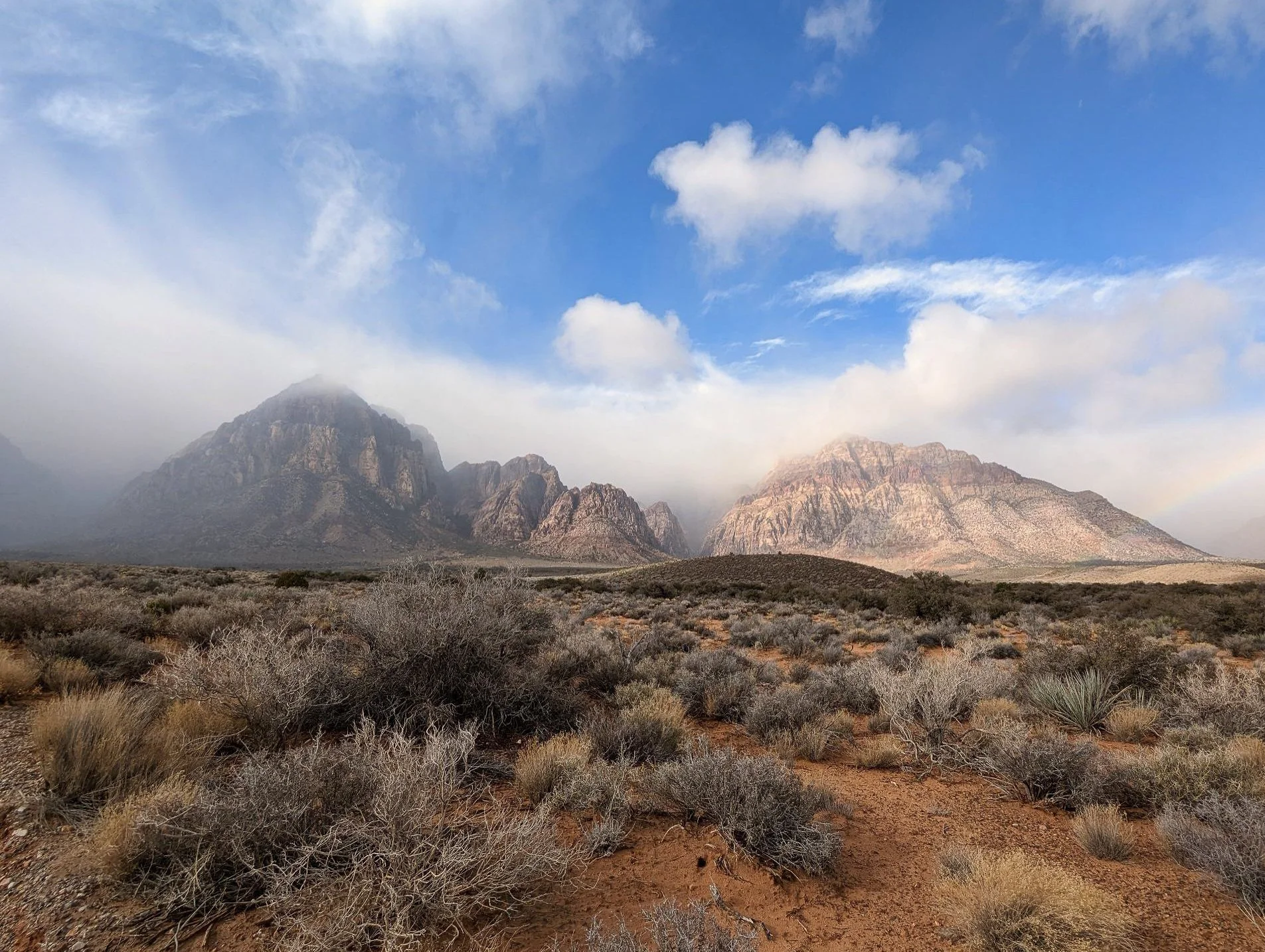 Desert landscape with shrubs and bushes in foreground, rugged mountains in background, partly cloudy sky with patches of blue, and a faint rainbow to the right.