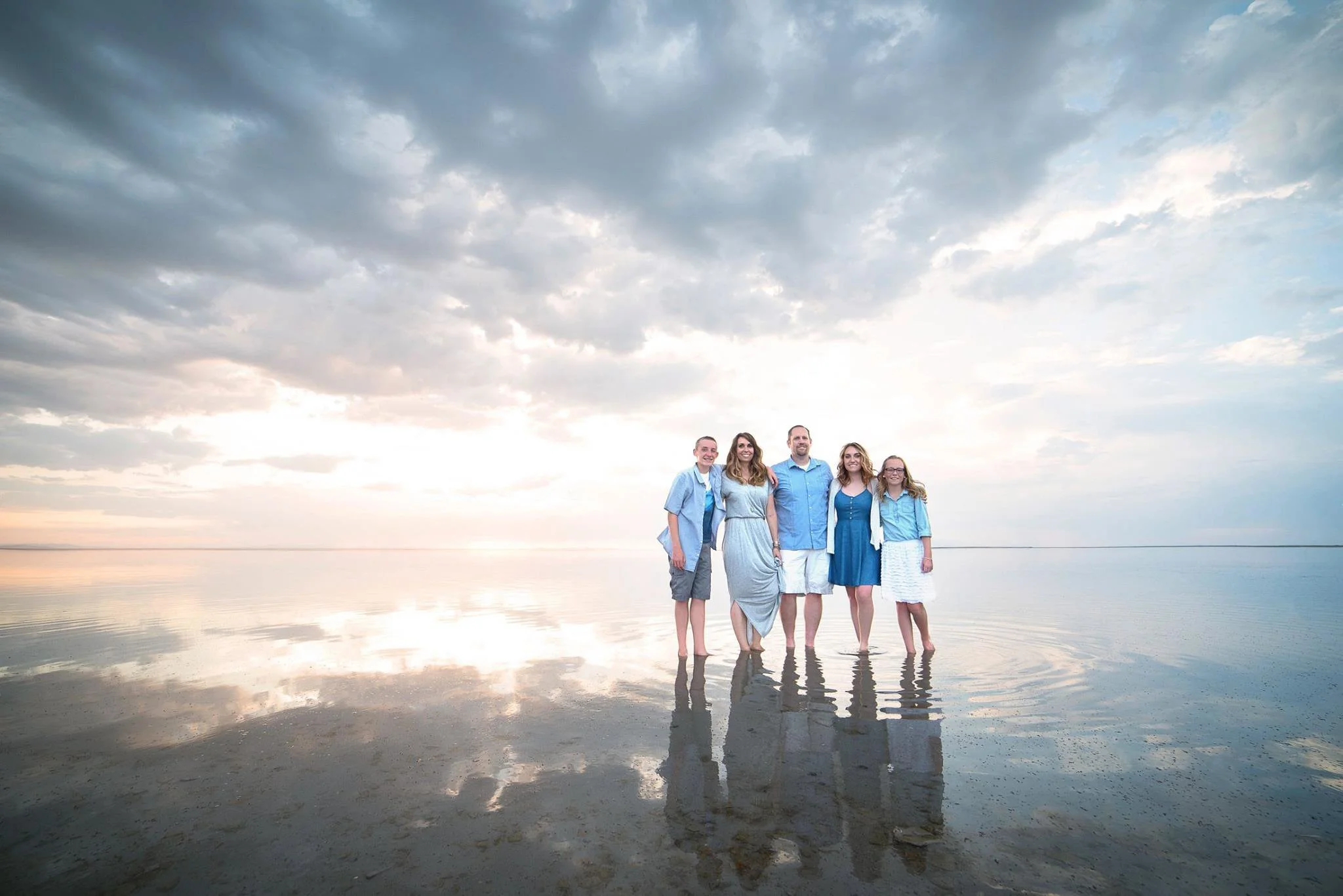 Group of five people standing in shallow water on a beach during sunset, with cloudy sky reflecting on the water.