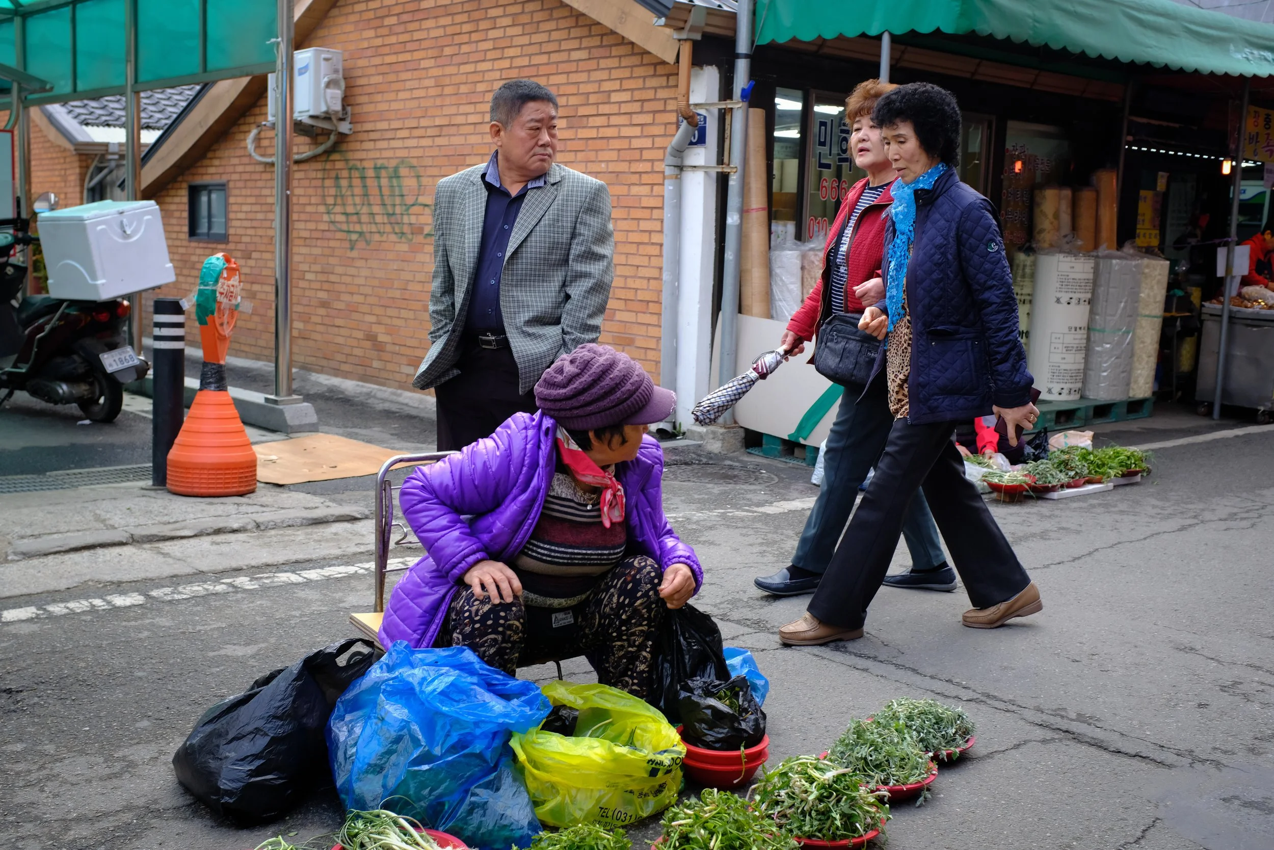 A street market scene with a woman in a purple jacket and hat sitting on a small stool, surrounded by plastic bags and potted plants. Three people are walking past her, and a brick wall with market stalls and a motorbike is visible in the background.