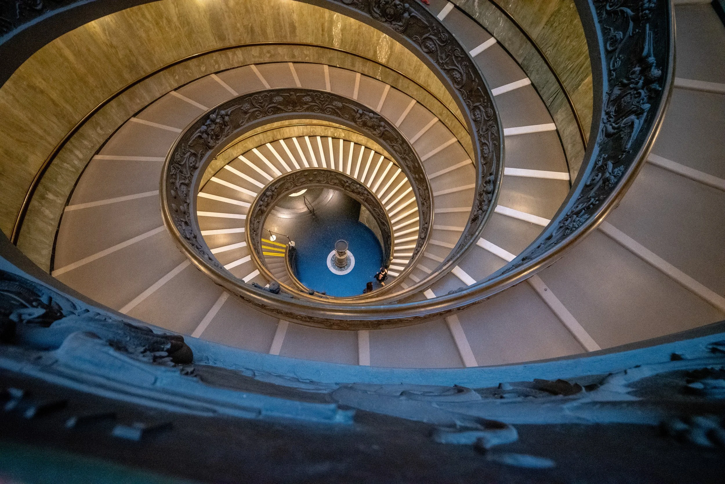 View looking down a spiraling staircase with ornate black railing, circular steps, and a blue floor at the bottom.