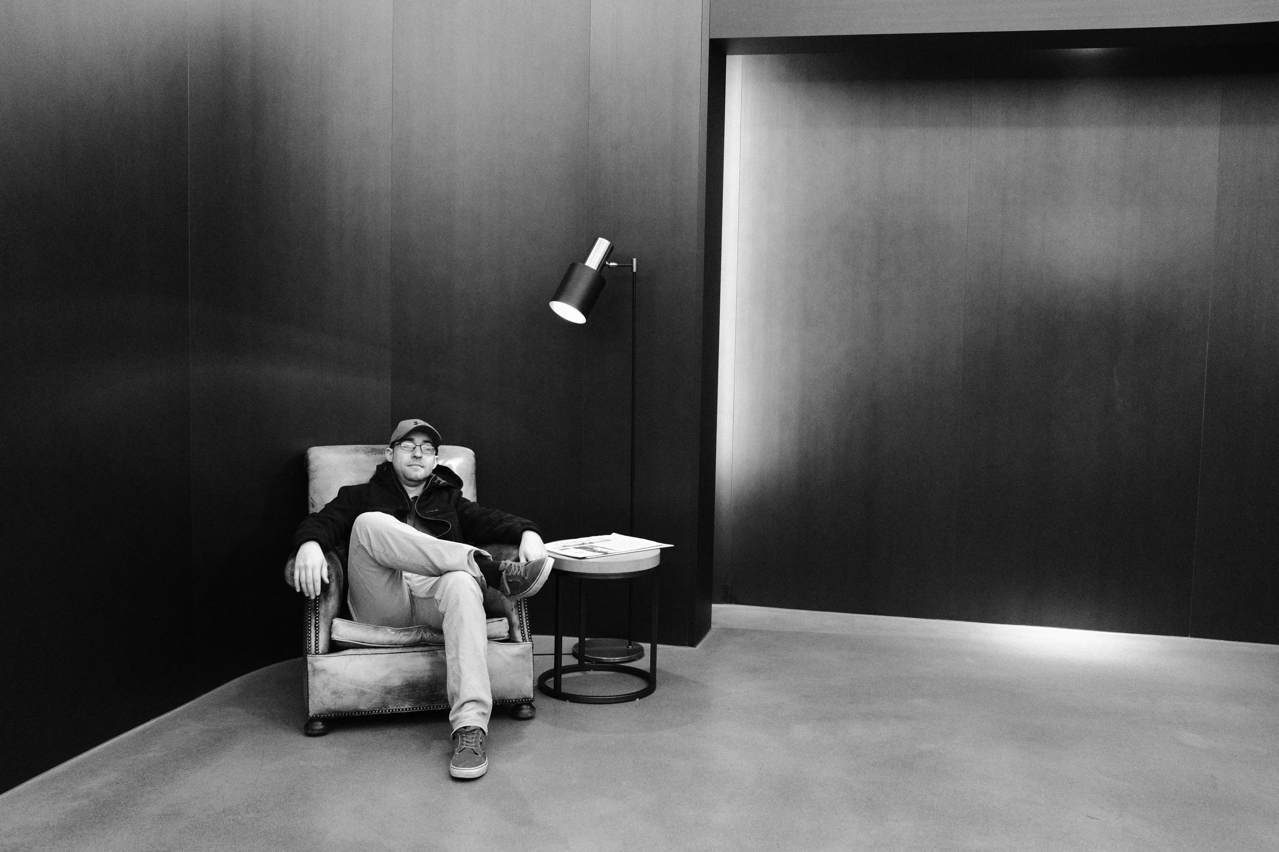 A man sitting comfortably on an armchair in a minimalist, modern interior with dark walls and a small side table next to him. The room features a floor lamp and an open large door in the background.
