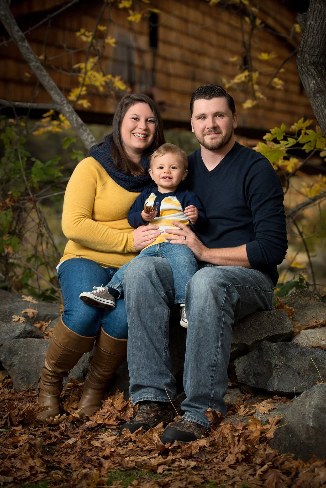 A family of three sitting outdoors on rocks surrounded by fallen leaves, with trees and a wooden fence in the background, during autumn.