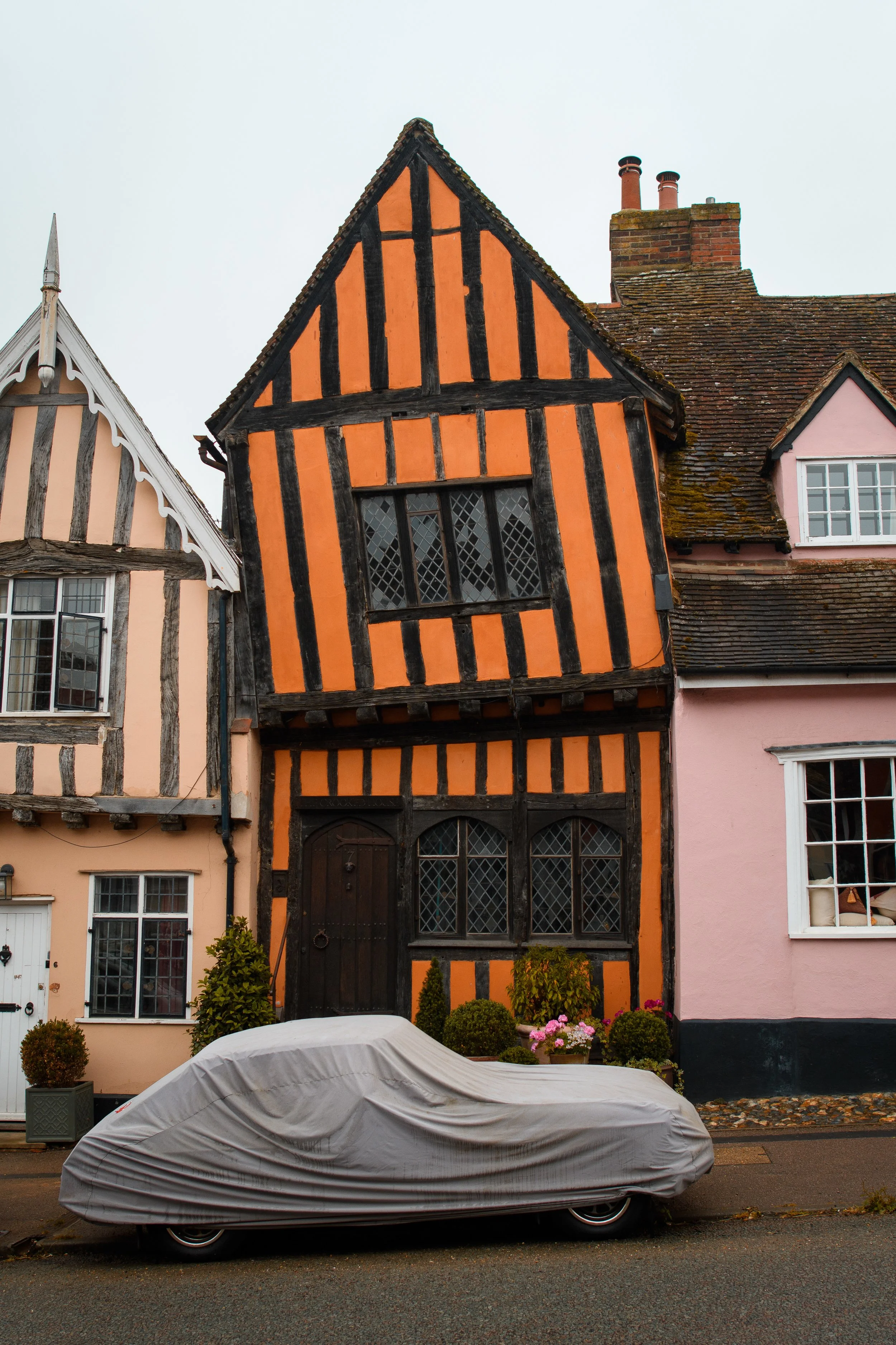 A row of colorful, historic Tudor-style houses with timber frames and leaded glass windows, with a parked car covered by a protective cover in front.