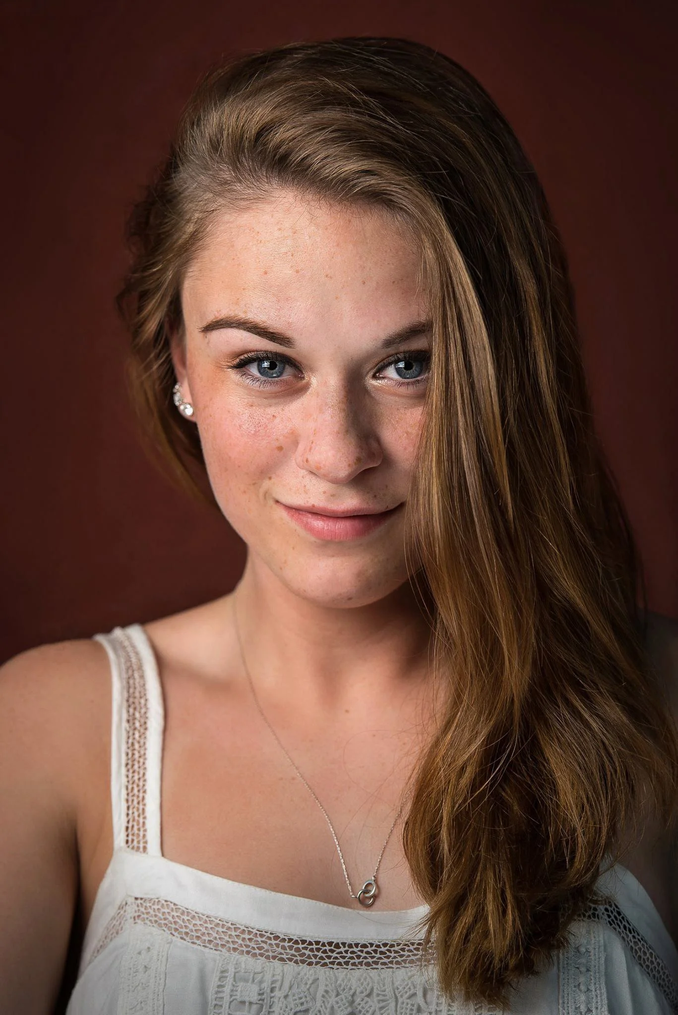 Close-up portrait of a young woman with long, wavy brown hair, blue eyes, and fair skin with freckles, wearing pearl earrings and a white top with lace details, against a dark brown background.