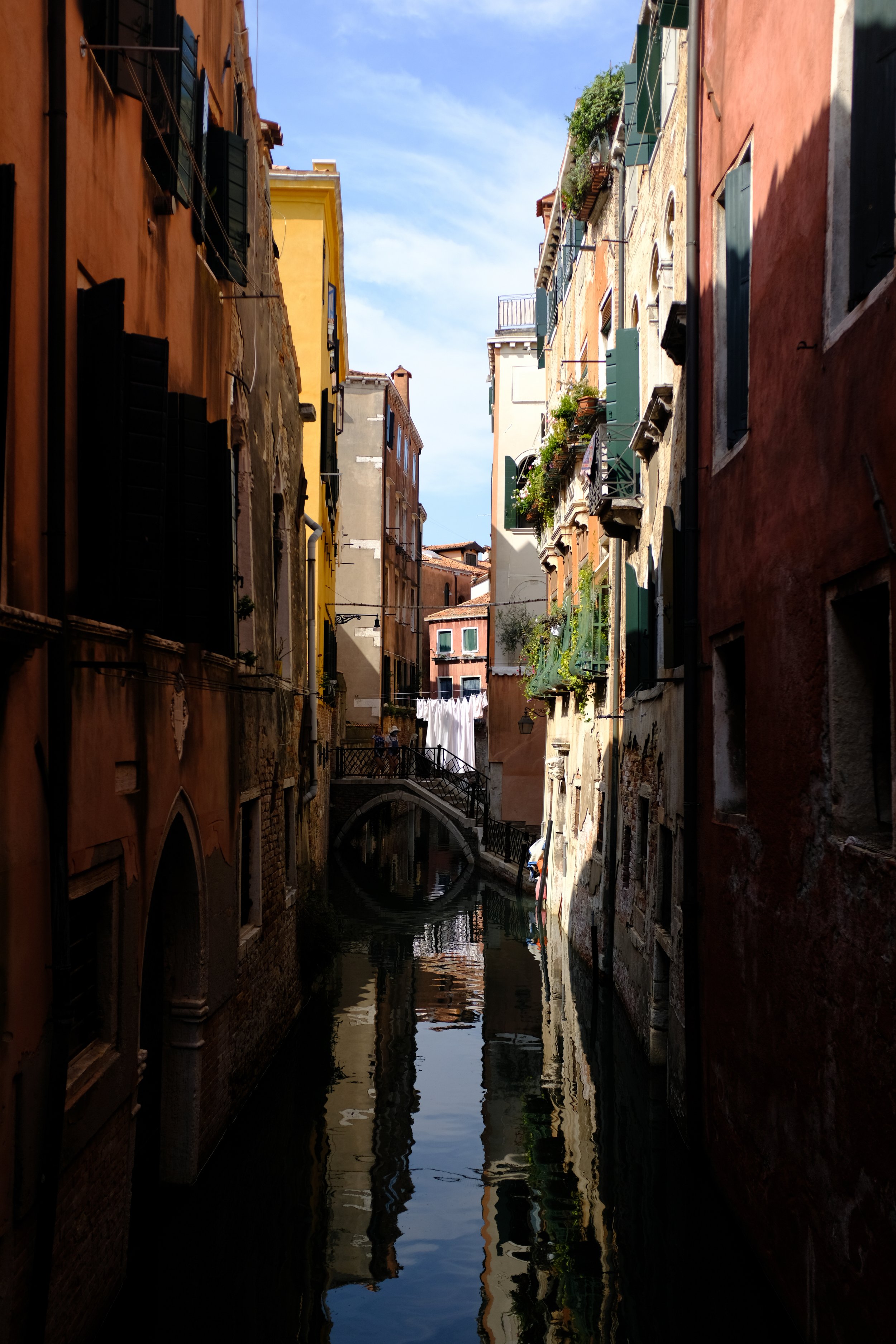 A narrow canal flanked by colorful, aged buildings with green shutters in Venice, Italy. A small footbridge crosses the canal, with laundry hanging to dry and a bright blue sky overhead.