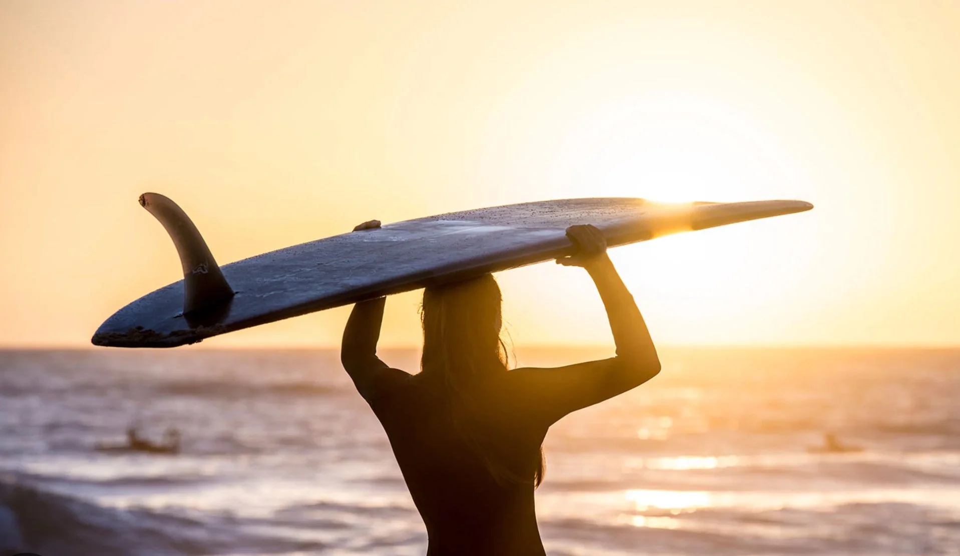 A person holding a surfboard over their head on a beach at sunset, with the ocean in the background.