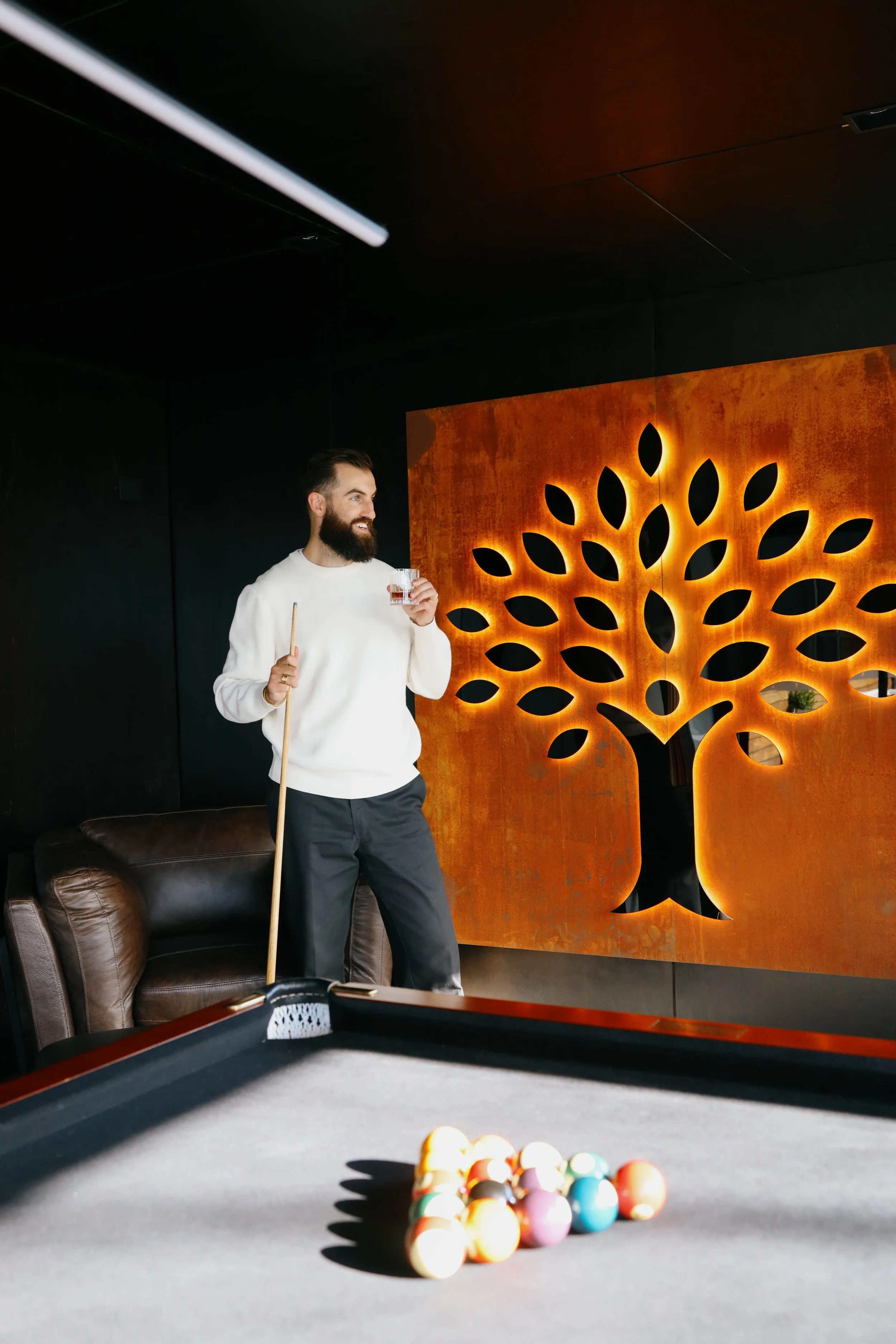 A man with a beard holding a pool cue and a glass, standing next to a brown leather chair beside a pool table with billiard balls racked, in a dimly lit room with a decorative orange wall featuring a backlit black floral pattern.