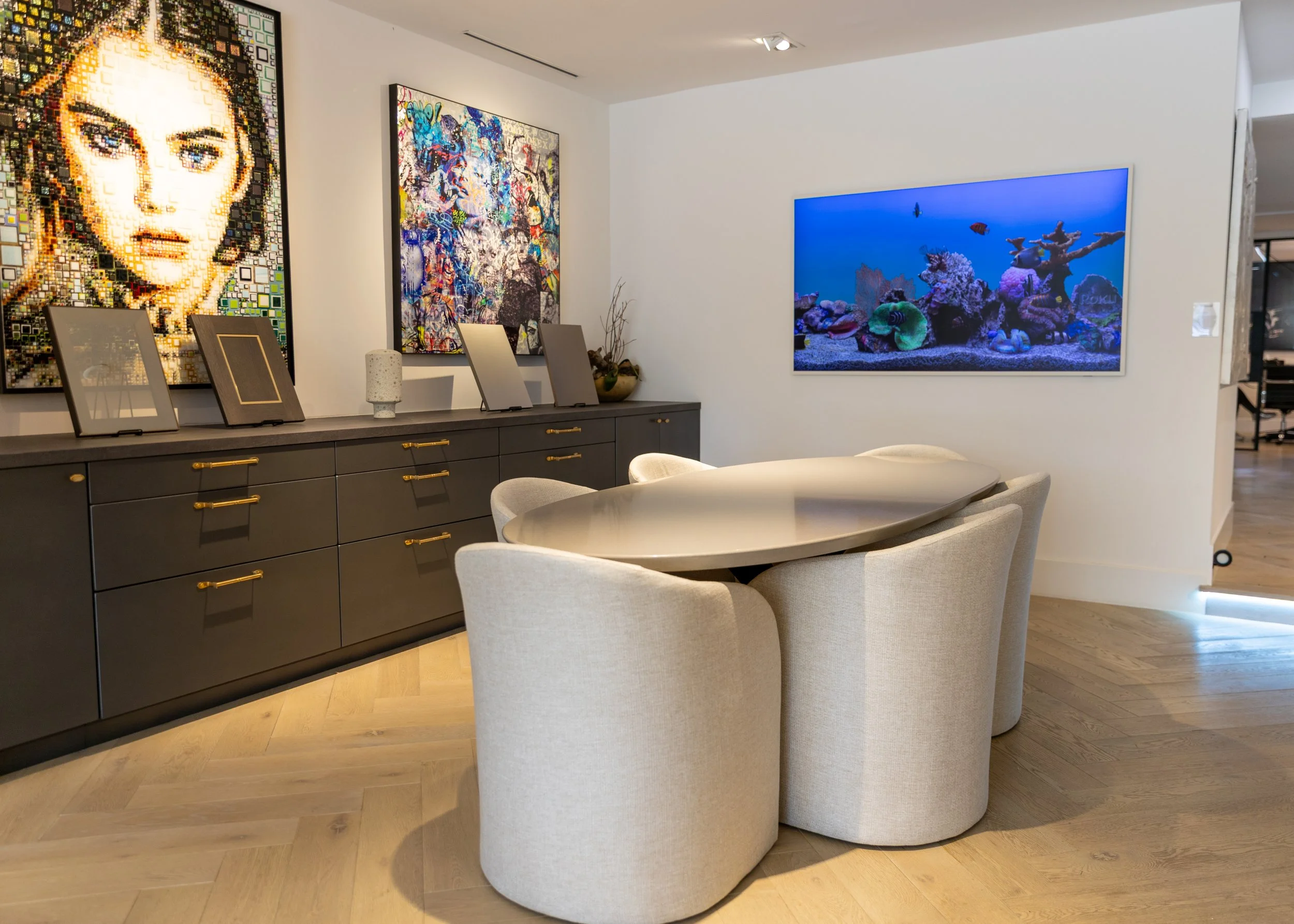 Modern dining room with a beige oval table and six matching upholstered chairs, dark gray sideboard with gold handles on the left, decorated with framed artworks and an art piece, and a large flat-screen TV displaying an underwater coral reef scene on the right wall.