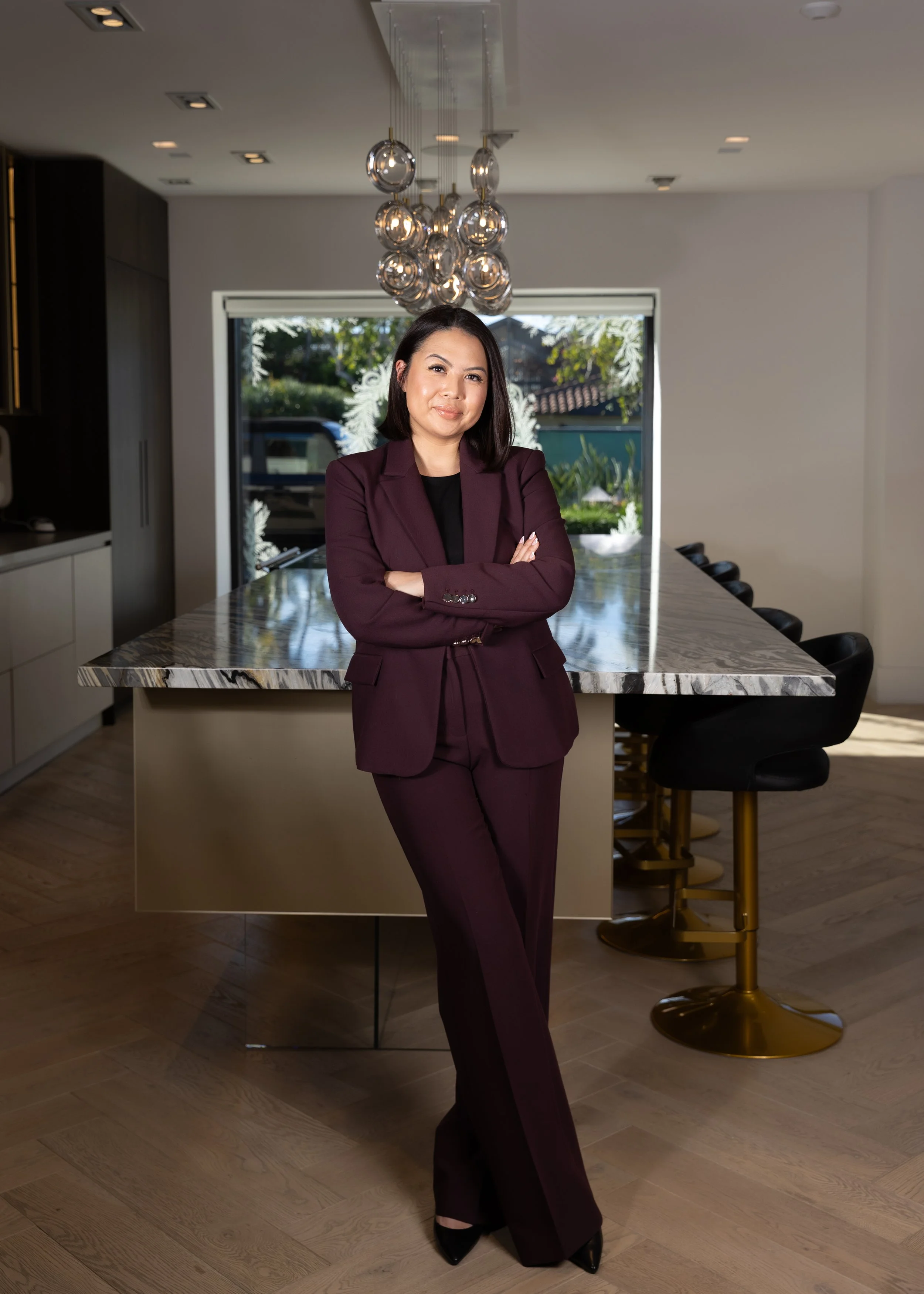 A woman in a burgundy suit standing with arms crossed in a modern kitchen with a marble island and bar stools, large window showing outdoor plants.