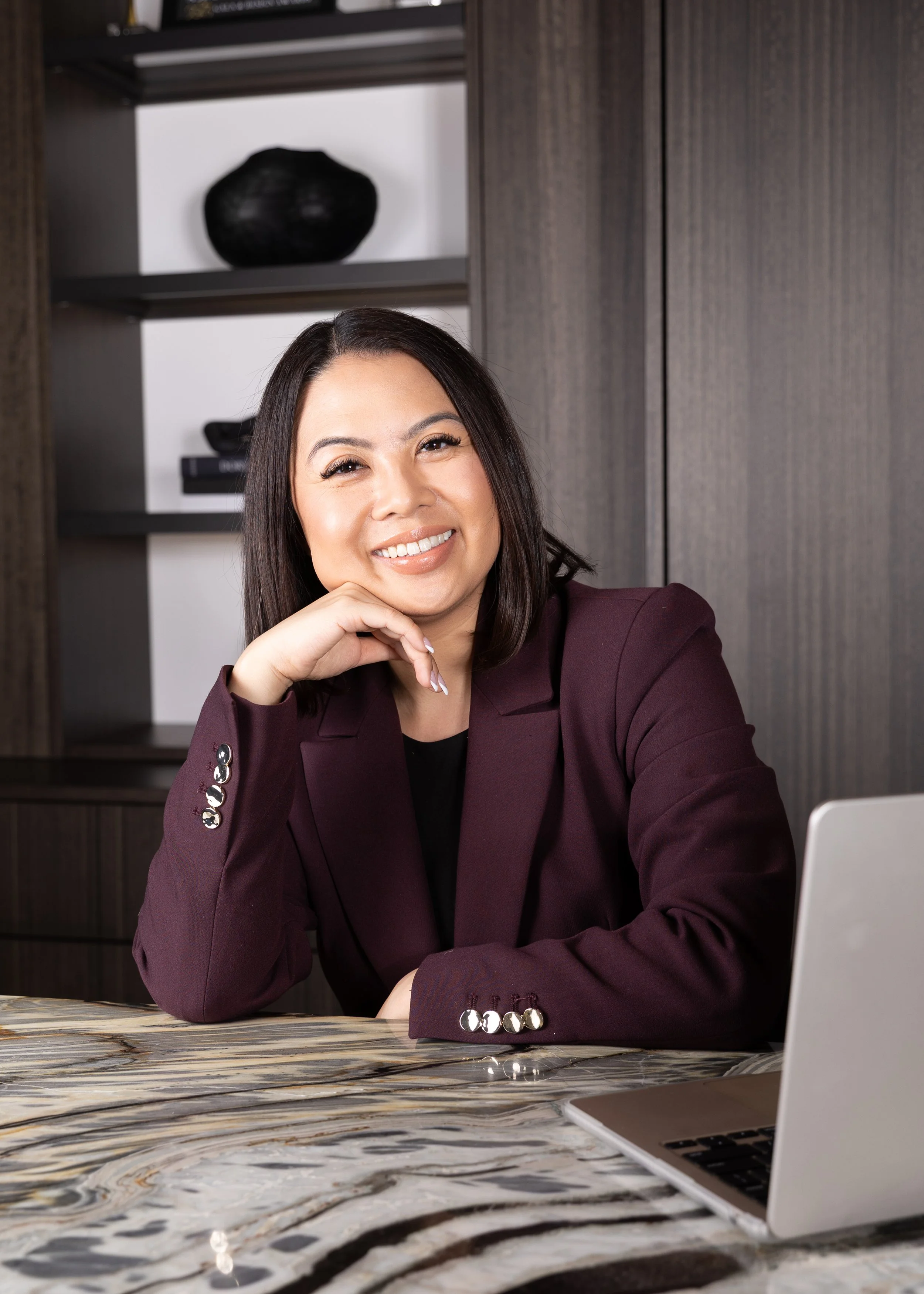 A woman with dark hair and a warm smile is sitting at a marble-topped table, resting her chin on her hand. She is wearing a maroon blazer with shiny buttons and a black top, and there is a laptop partially visible in front of her. The background shows a dark wooden cabinet with shelves and a decorative black vase.