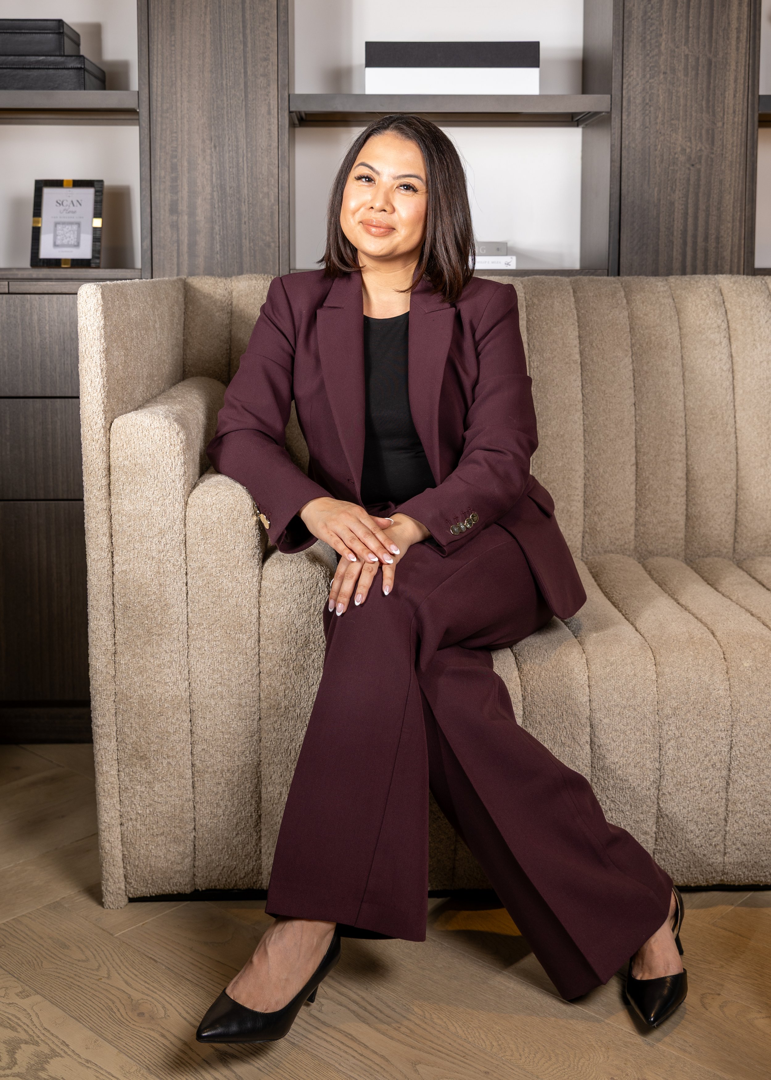 A woman in a burgundy suit sitting on a beige couch in an office setting.
