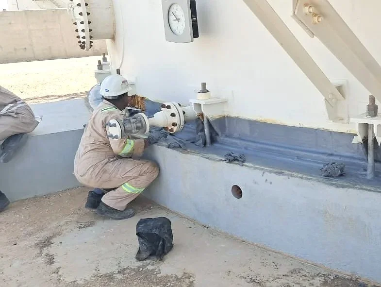 A worker in safety gear inspecting or working on a large industrial tank outside. The worker is wearing a hard hat, gloves, and coveralls, and is bent down near the tank bottom which is protected using the Trenton Anti-Corrosion wax tape Wrap system.