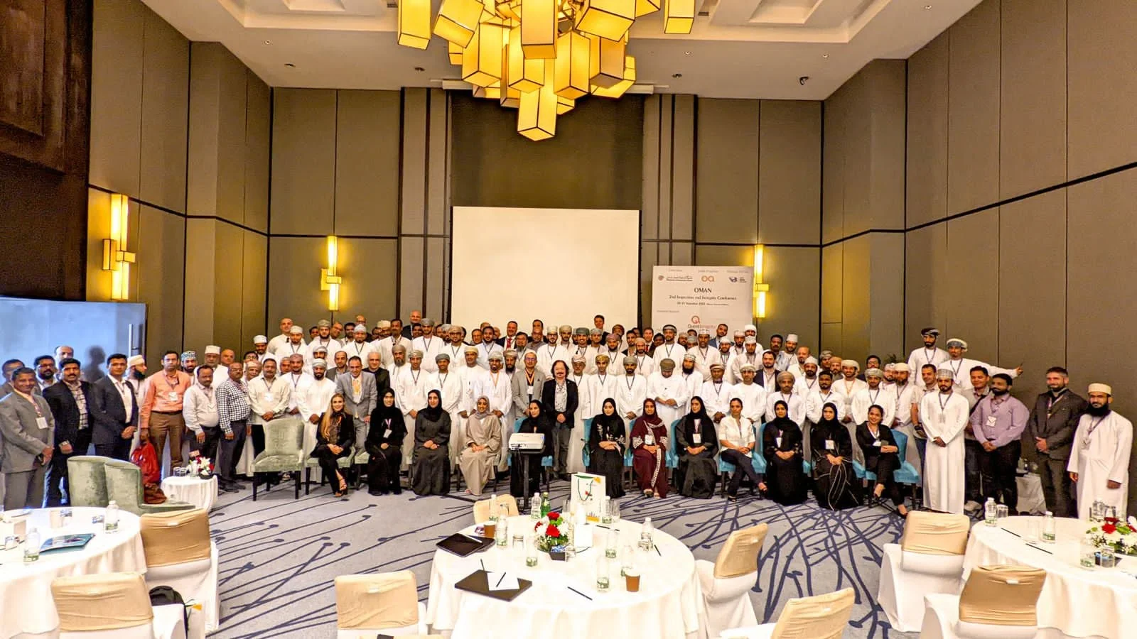 Group photo of conference attendees in a large banquet hall with high ceilings, warm lighting, and round tables with water bottles and notepads.