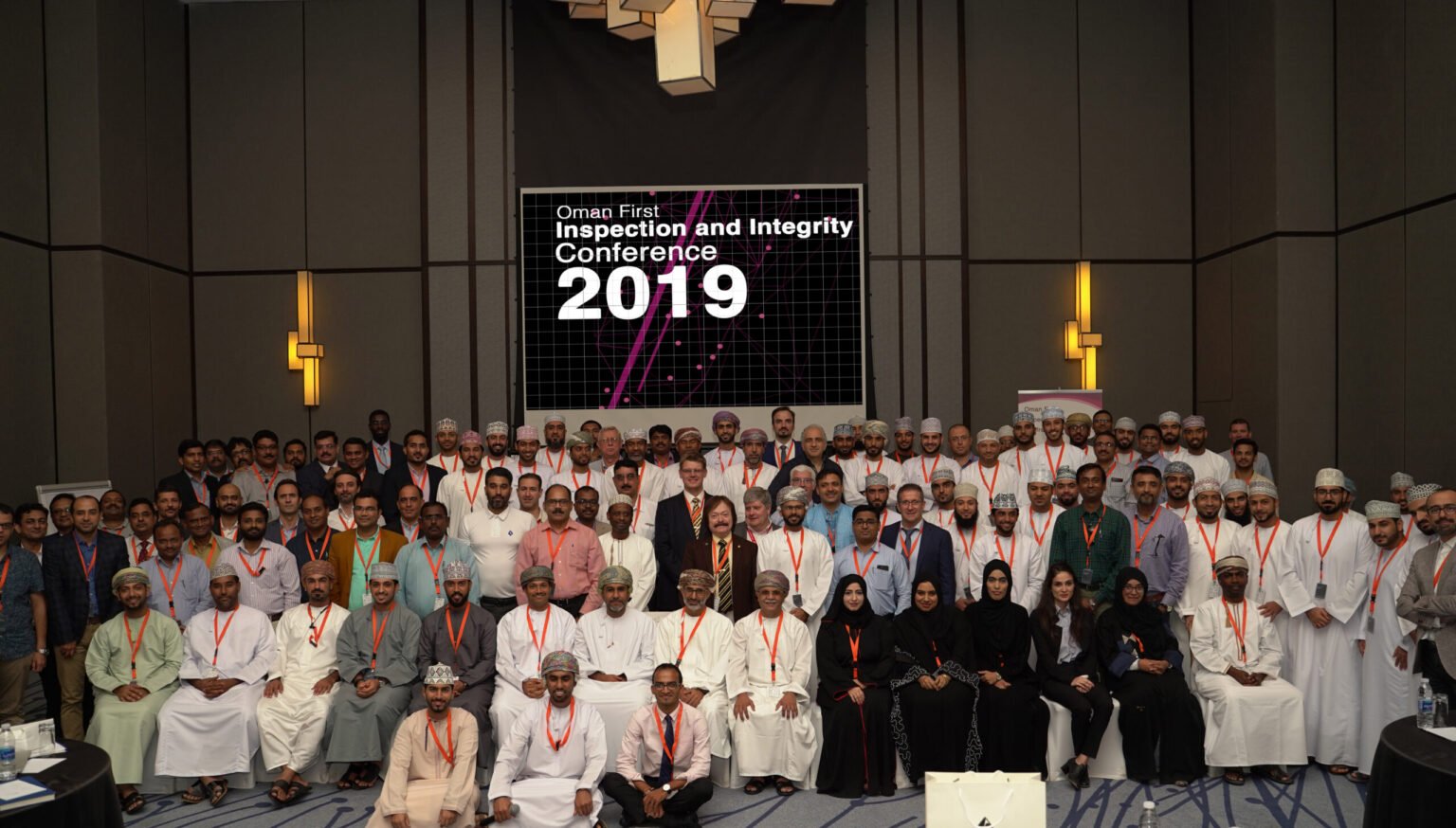 Group photo of conference attendees at the Oman First Inspection and Integrity Conference 2019, with a large screen displaying the conference details in the background.