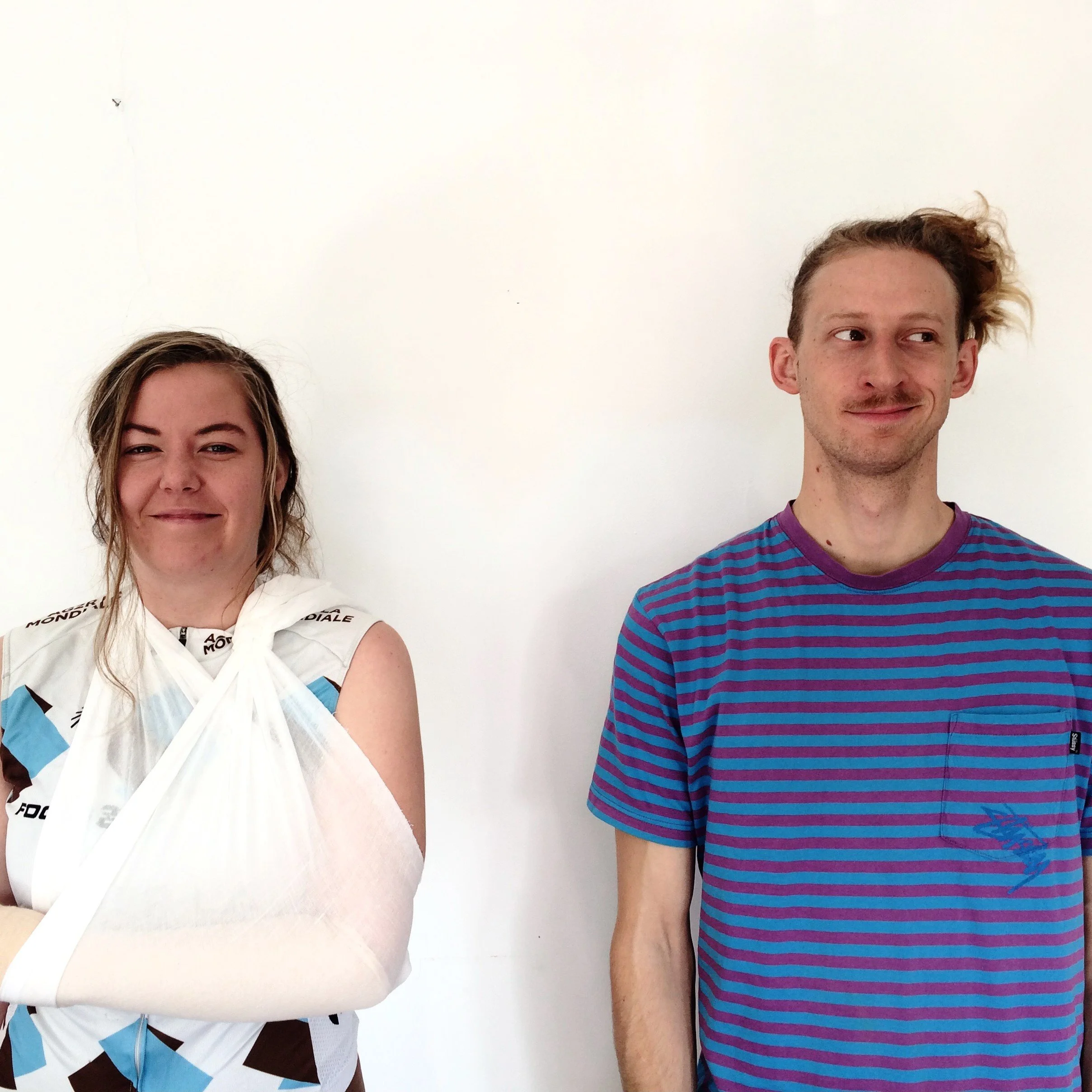 A woman and a man stand next to each other facing the camera. We see them from the wait up. She is on the left looking directly at us, smiling with her arm in a sling. He looks off the the right with a smirk wearing a colourful t-shirt.