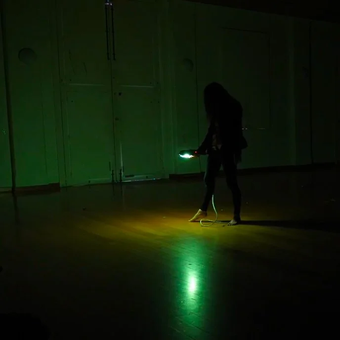 Two figures dressed in black dance in the distance of a large room. One of them holds an eerie green lamp, which is the only source of light. The walls are dirty and white with some large white doors in the background. There are wooden floorboards.