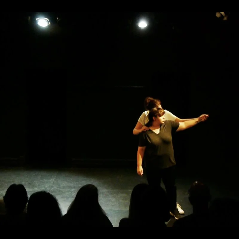 Two women stand on a bare stage in front of an audience, one behind the other. The one in front wear black pants and a t-shirt. The one behind wears a white t-shirt and holds the other woman's arm extended out to the left by her hand.