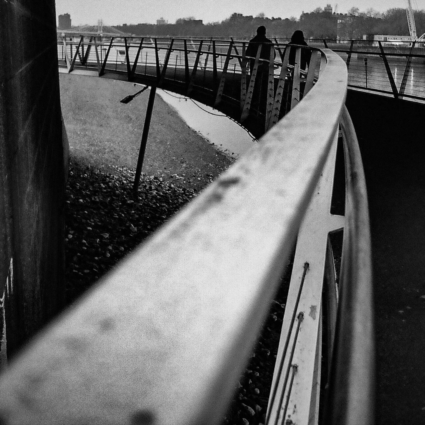 Black and white photo of a curving pedestrian bridge with two people walking on it, seen from a low angle.