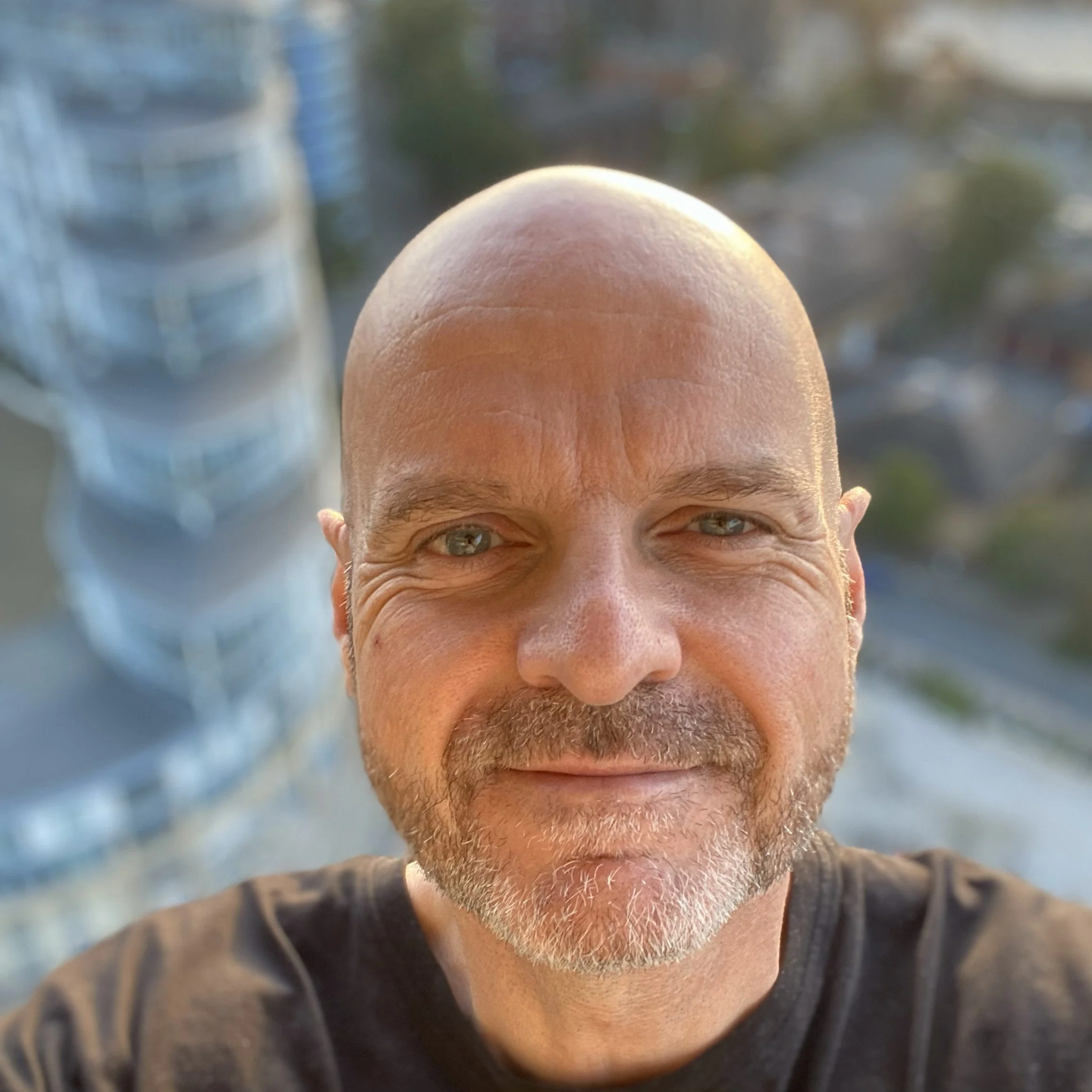 Close-up of a bald man with a beard smiling, standing outdoors with tall buildings in the background.