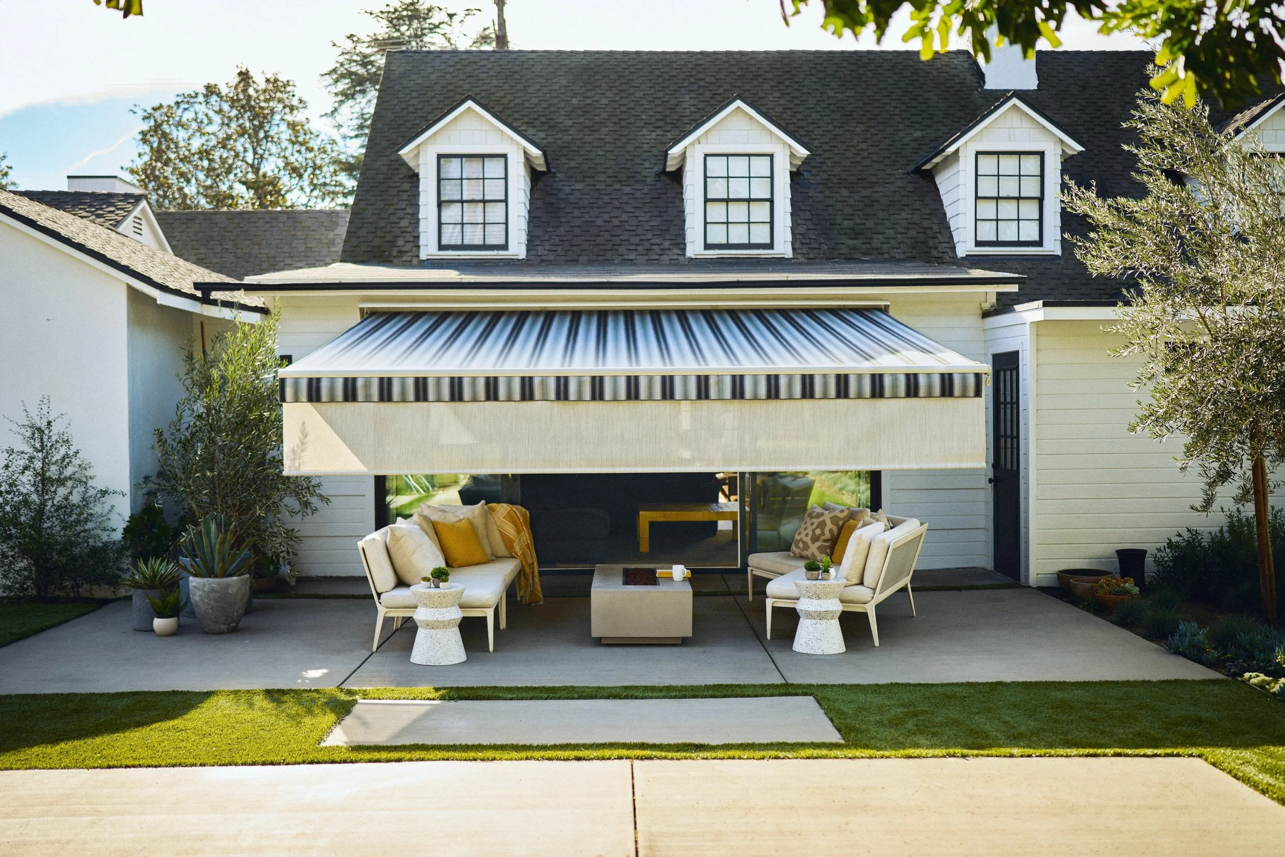 Backyard patio with outdoor furniture, potted plants, and a house with three dormer windows and an awning.