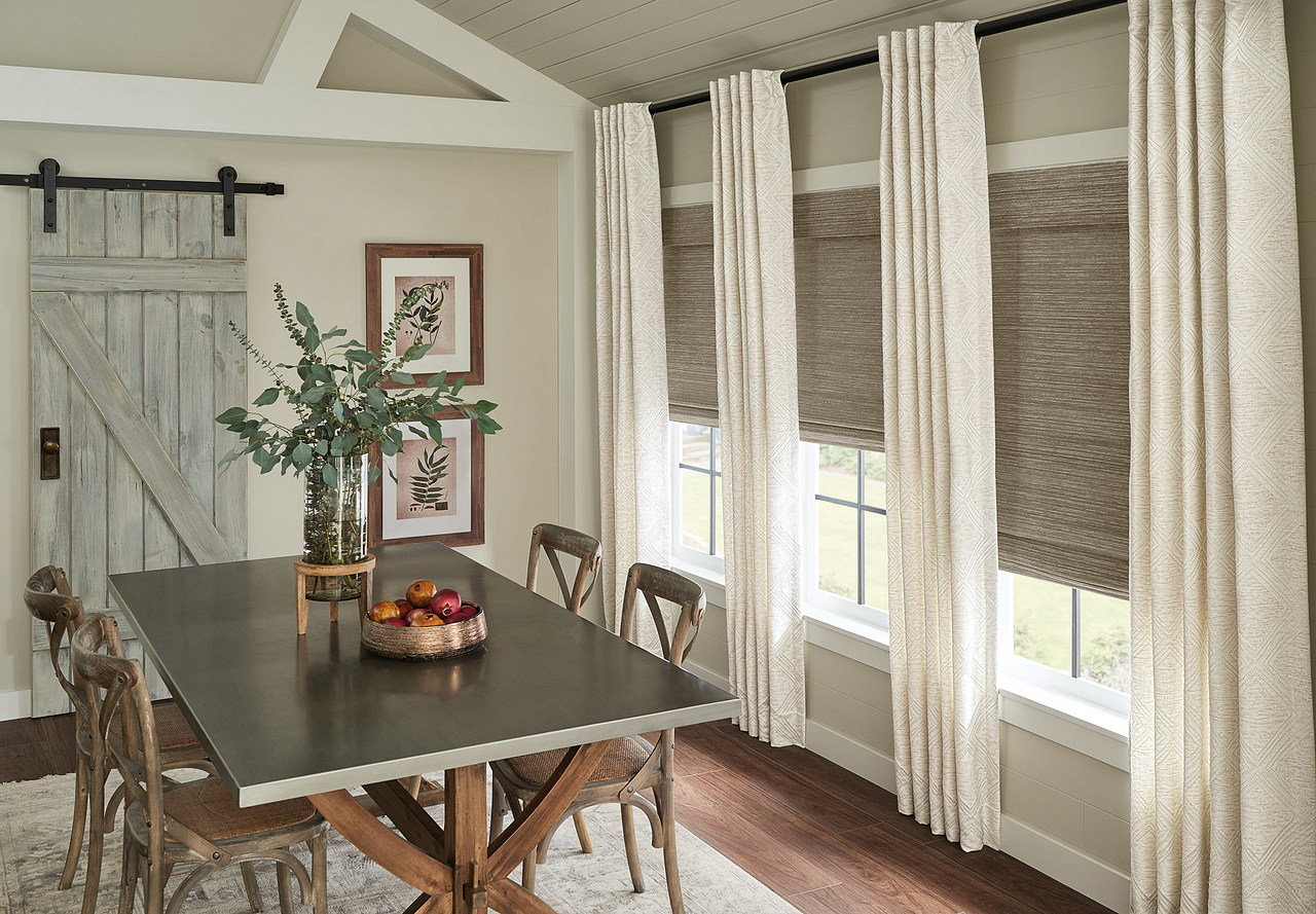 Dining room with a dark wood table, six wooden chairs, a vase with green foliage, framed botanical prints on the wall, a sliding barn door, and large windows with light curtains and dark shades.