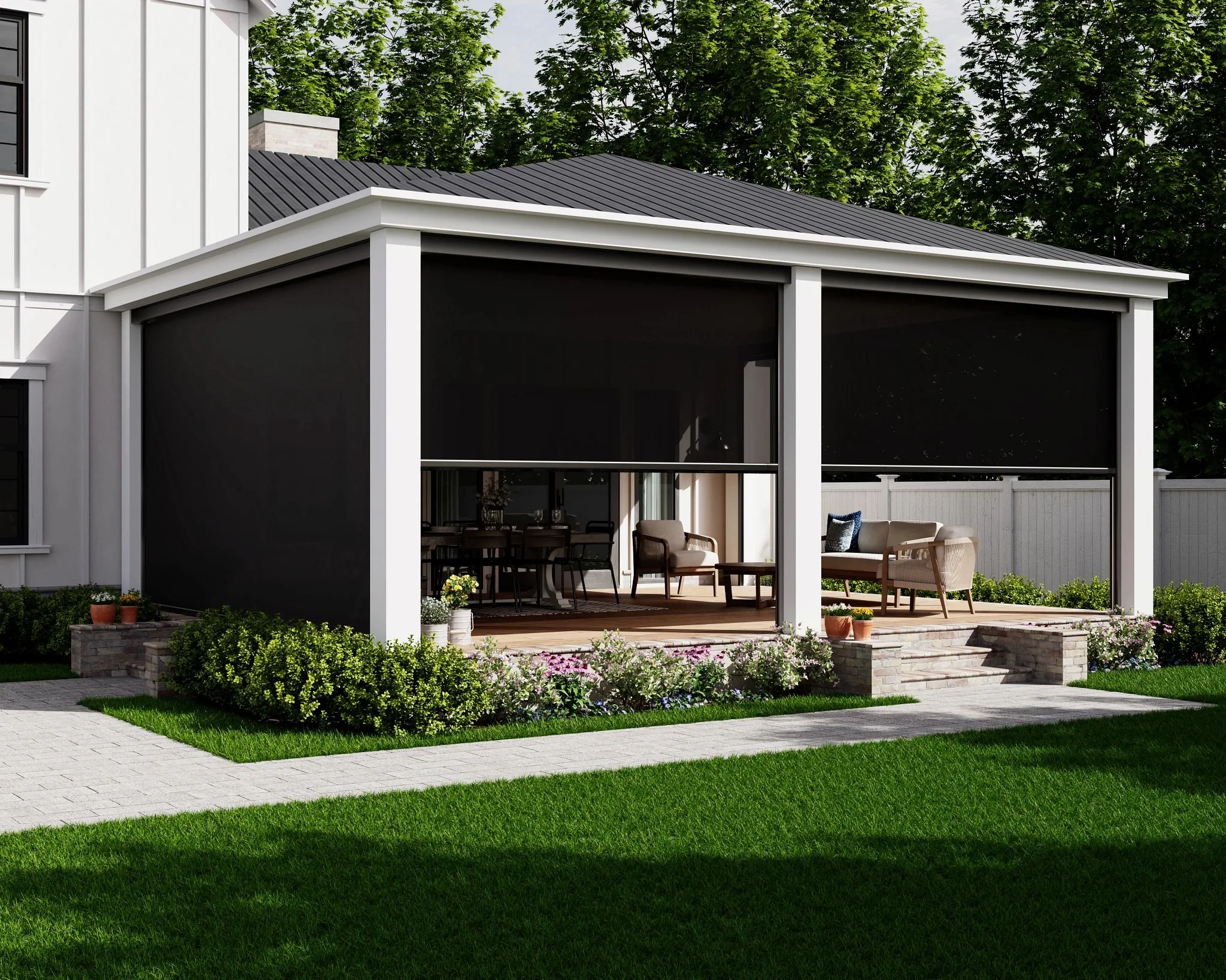 The image shows a modern house exterior with a covered outdoor patio. The house has white siding, a dark metal roof, and large sliding glass doors. The patio features outdoor seating with beige cushions and small tables. There are green bushes and trees in the background, along with a well-maintained lawn and stone pathway.