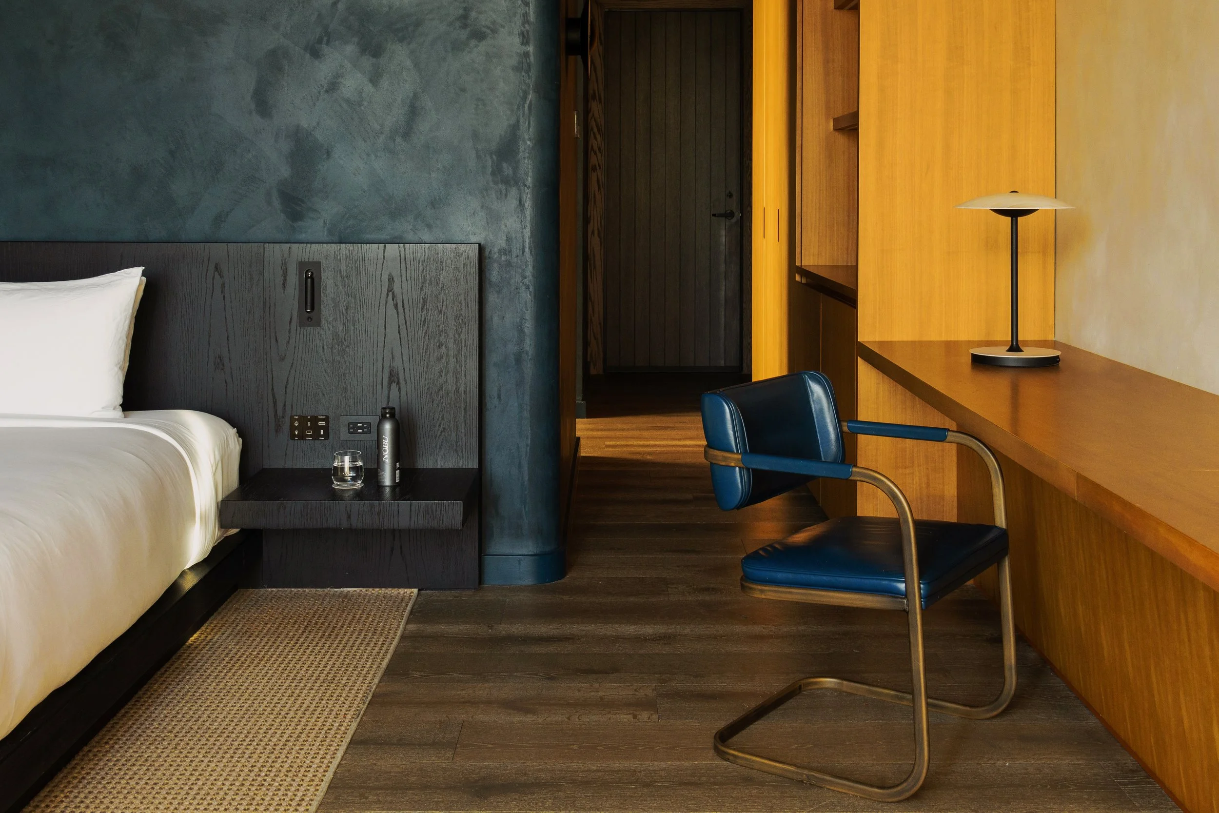 Interior of a modern hotel room with a bed, a nightstand with a water bottle and glass, a navy blue desk chair, a wooden desk with a lamp, and a textured wall in the background.