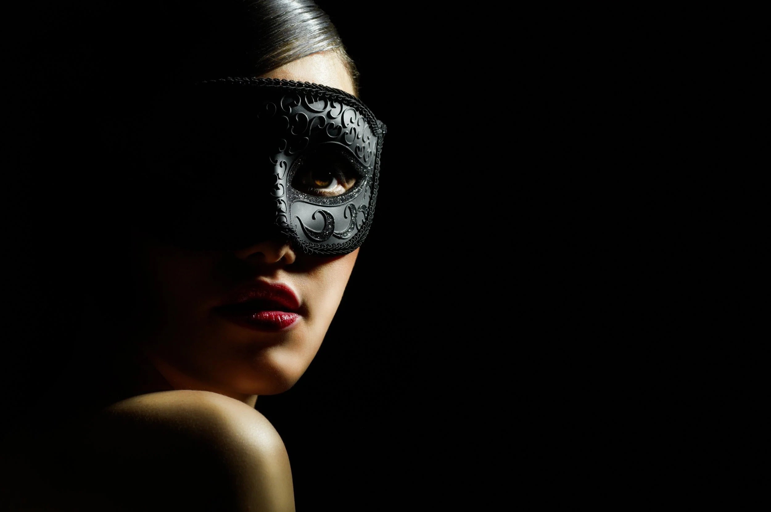 A woman with dark lipstick and hair, wearing an ornate black mask covering one eye, against a black background.