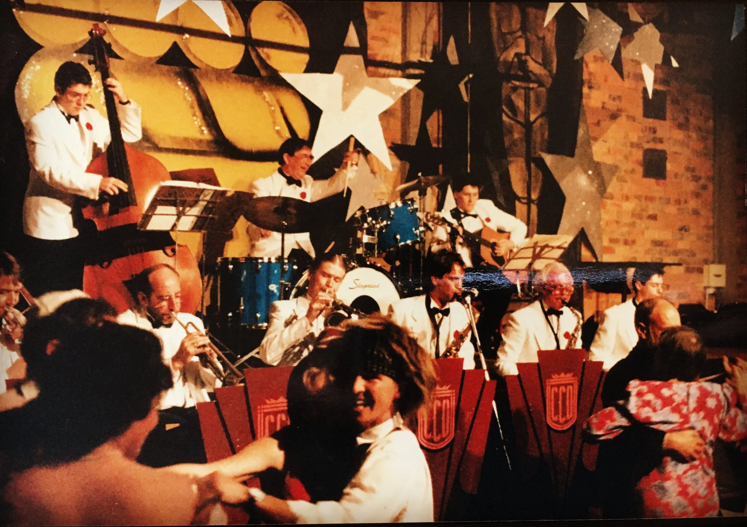 Big band jazz orchestra performing on stage with a backdrop of large stars and musical notes, dancers in front of the stage.