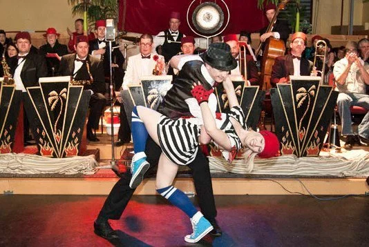 Two performers dancing in front of a seated orchestra with musicians in formal attire, playing instruments, in an indoor venue.