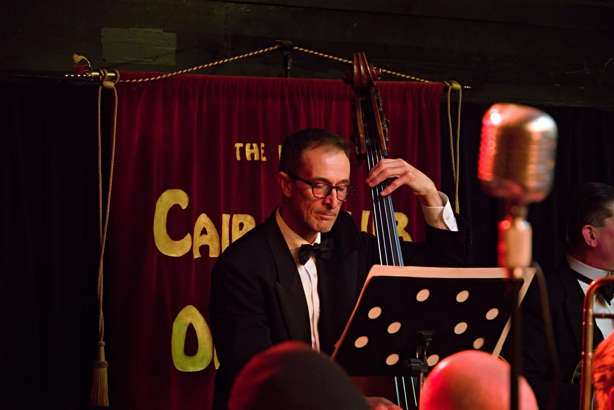 A jazz musician in a black tuxedo and bow tie playing a double bass on stage with a red curtain backdrop. The scene is dimly lit, and a vintage microphone is visible in the foreground. The backdrop has yellow letters spelling out "The Capital of."