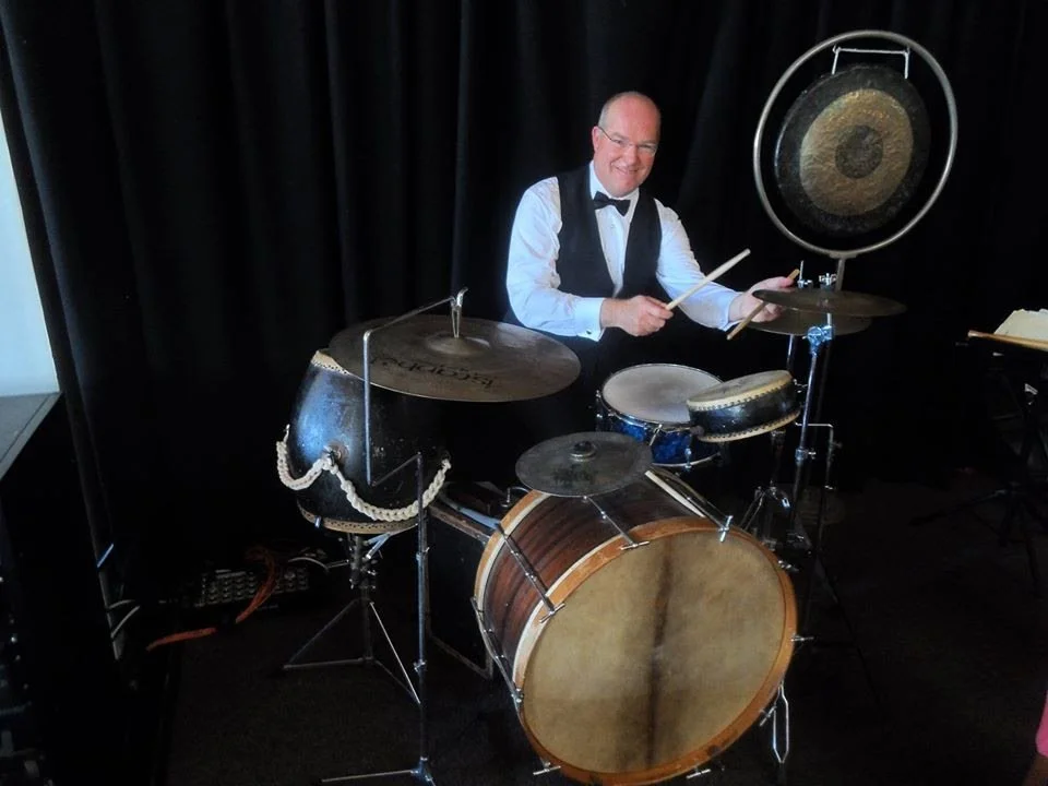 A man in a tuxedo vest and bow tie is playing a drum set, smiling at the camera, in front of a black curtain.