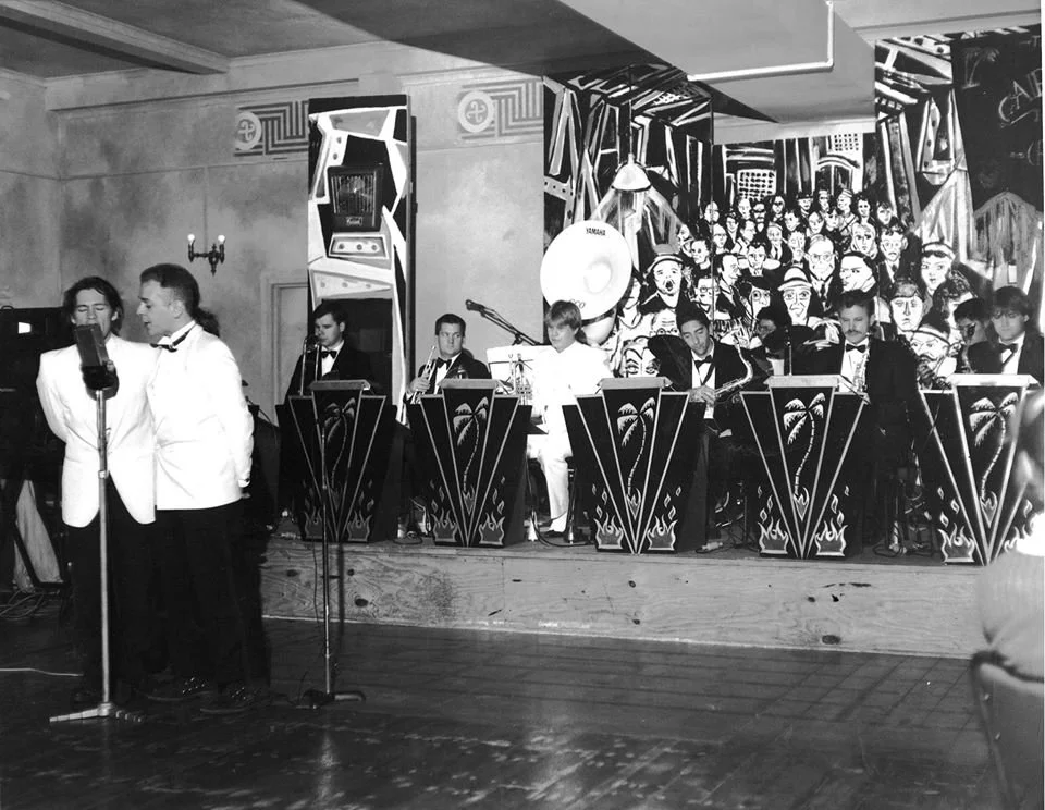 A black and white photo of a musical performance with several people, some dressed in tuxedos, playing instruments and singing on stage. There is a mural of people in the background.
