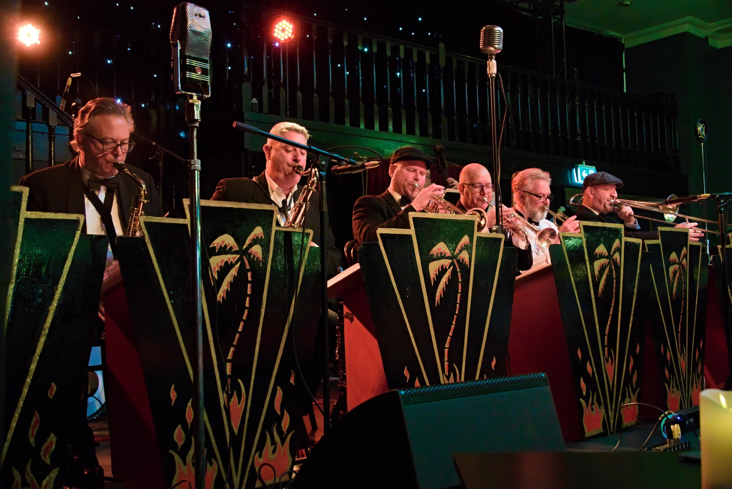 A jazz band with seven musicians playing horn instruments on stage, decorated with palm tree designs on music stands, under stage lights.